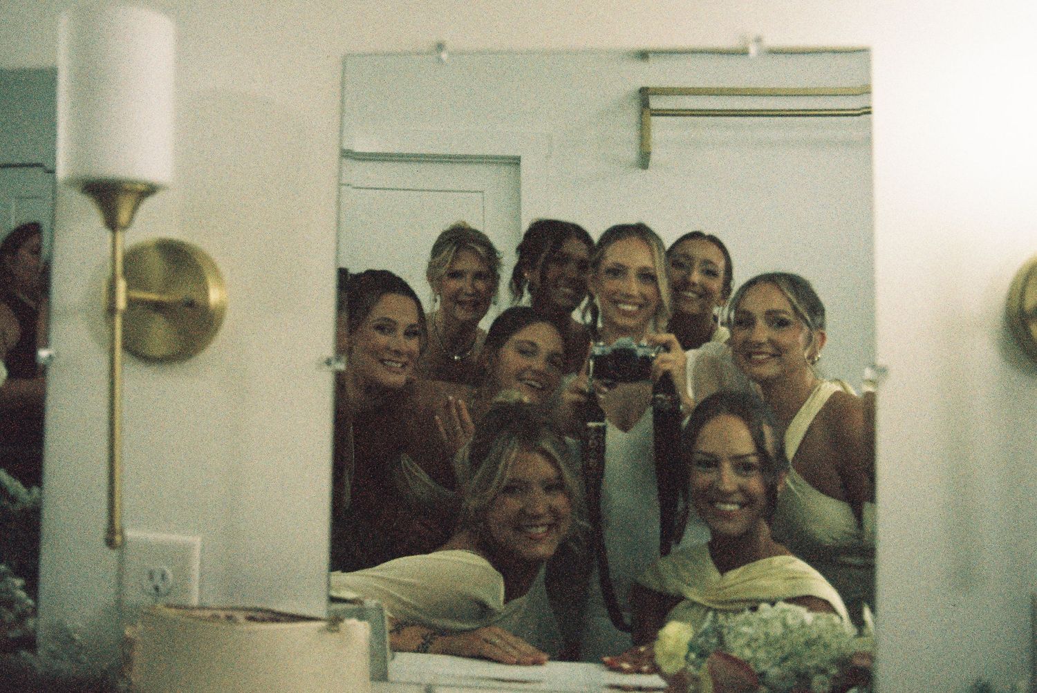 Group of bridesmaids smiling together in mirror selfie, wearing neutral tones, holding bouquet in elegant dressing room.