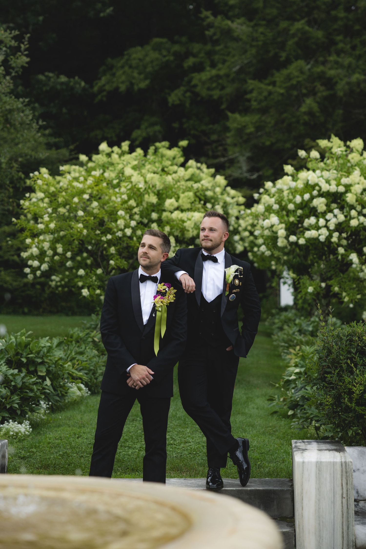 Two grooms in black tuxedos with boutonnieres pose together in a lush garden with white flowering bushes.