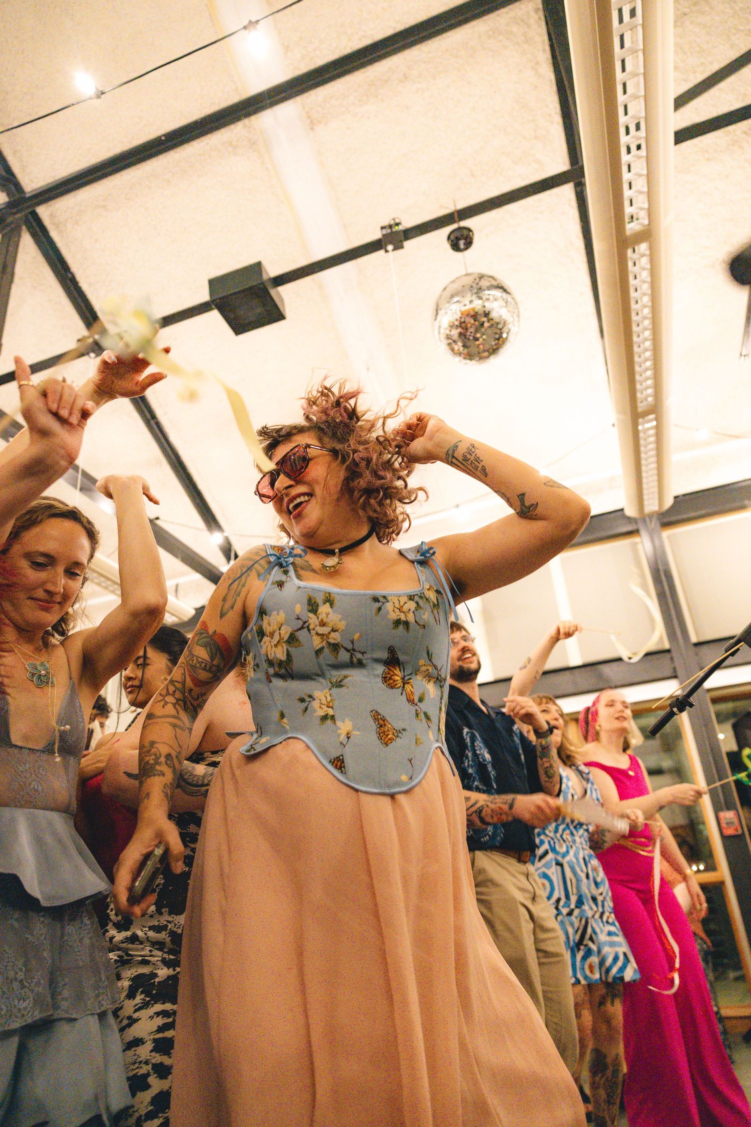 Tattooed woman in floral dress dancing joyfully under disco ball at lively party celebration.