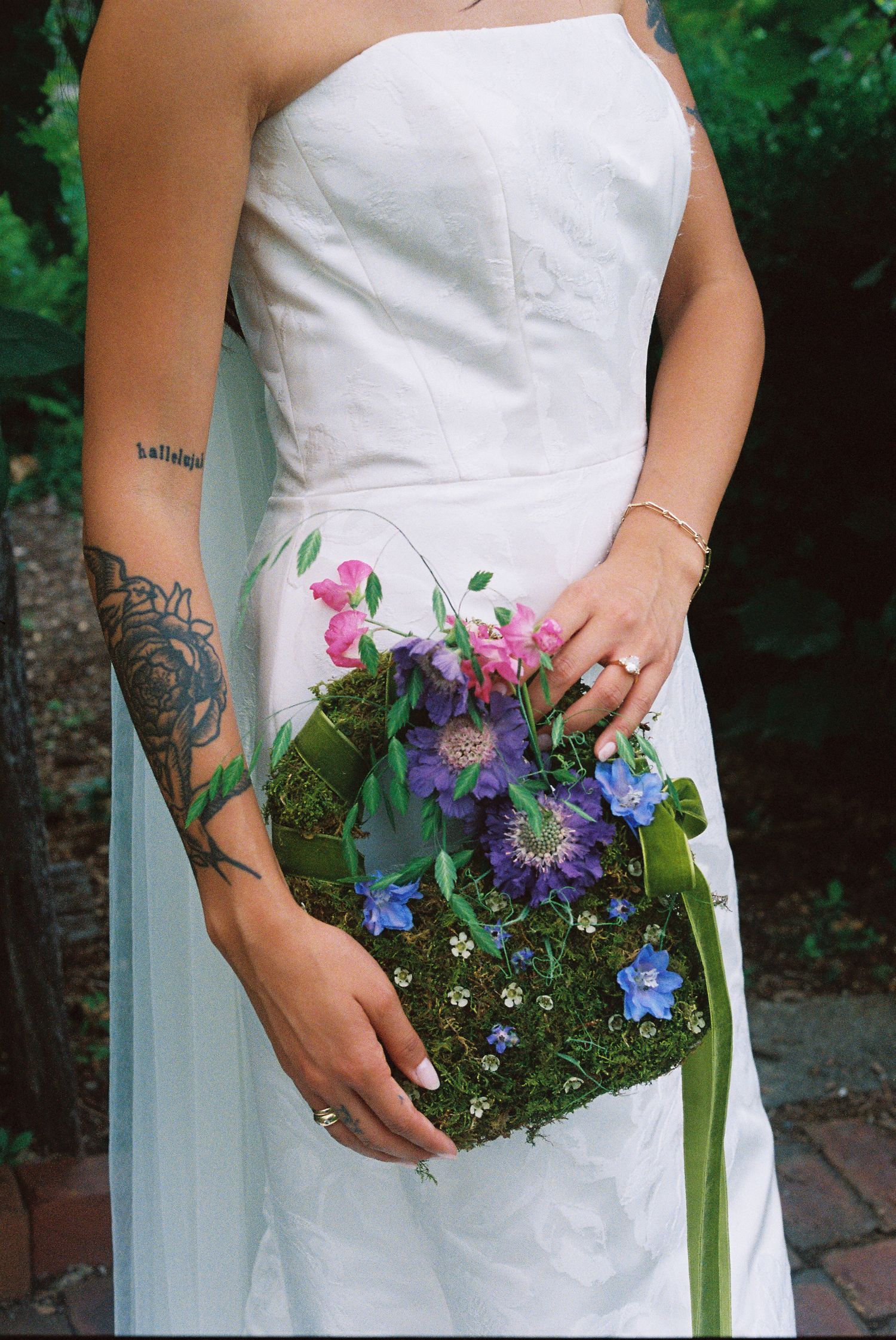 Tattooed bride in white strapless gown holds a moss ball bouquet with purple, blue, and pink wildflowers.