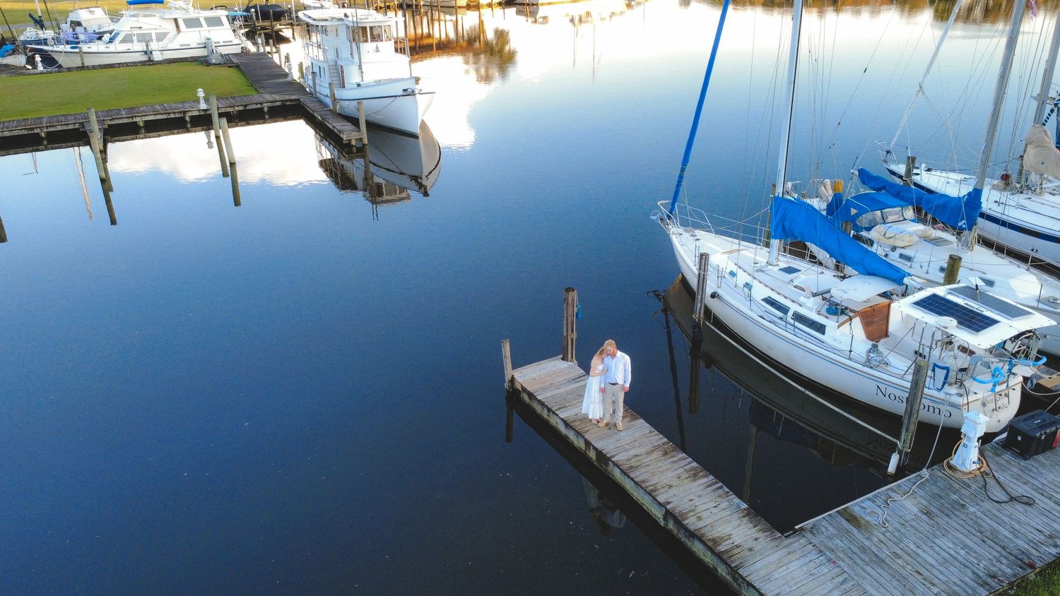Aerial view of a couple standing on a wooden dock surrounded by sailboats in a calm marina at golden hour.