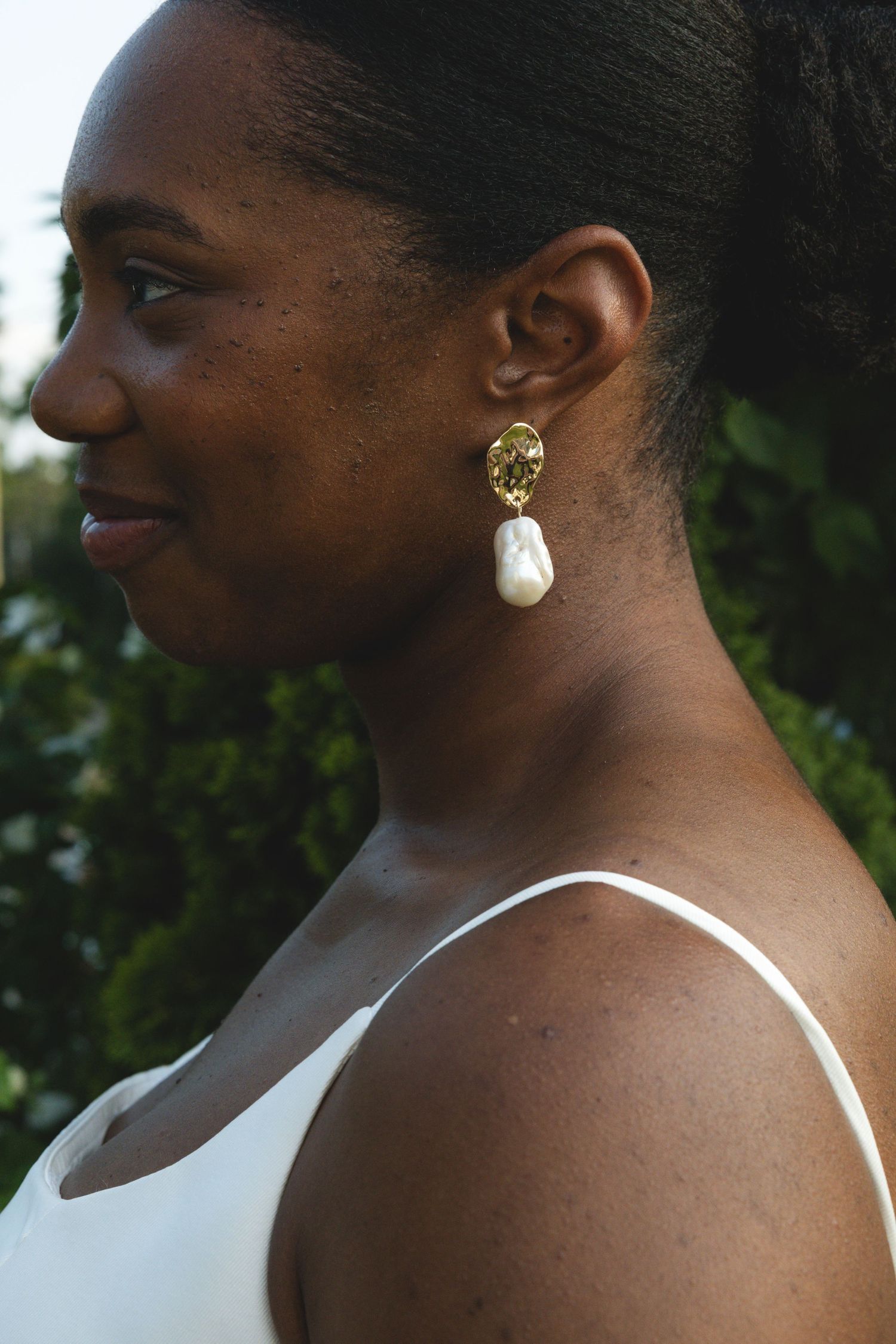 Woman wearing elegant gold and baroque pearl drop earrings, smiling in profile view, dressed in white.