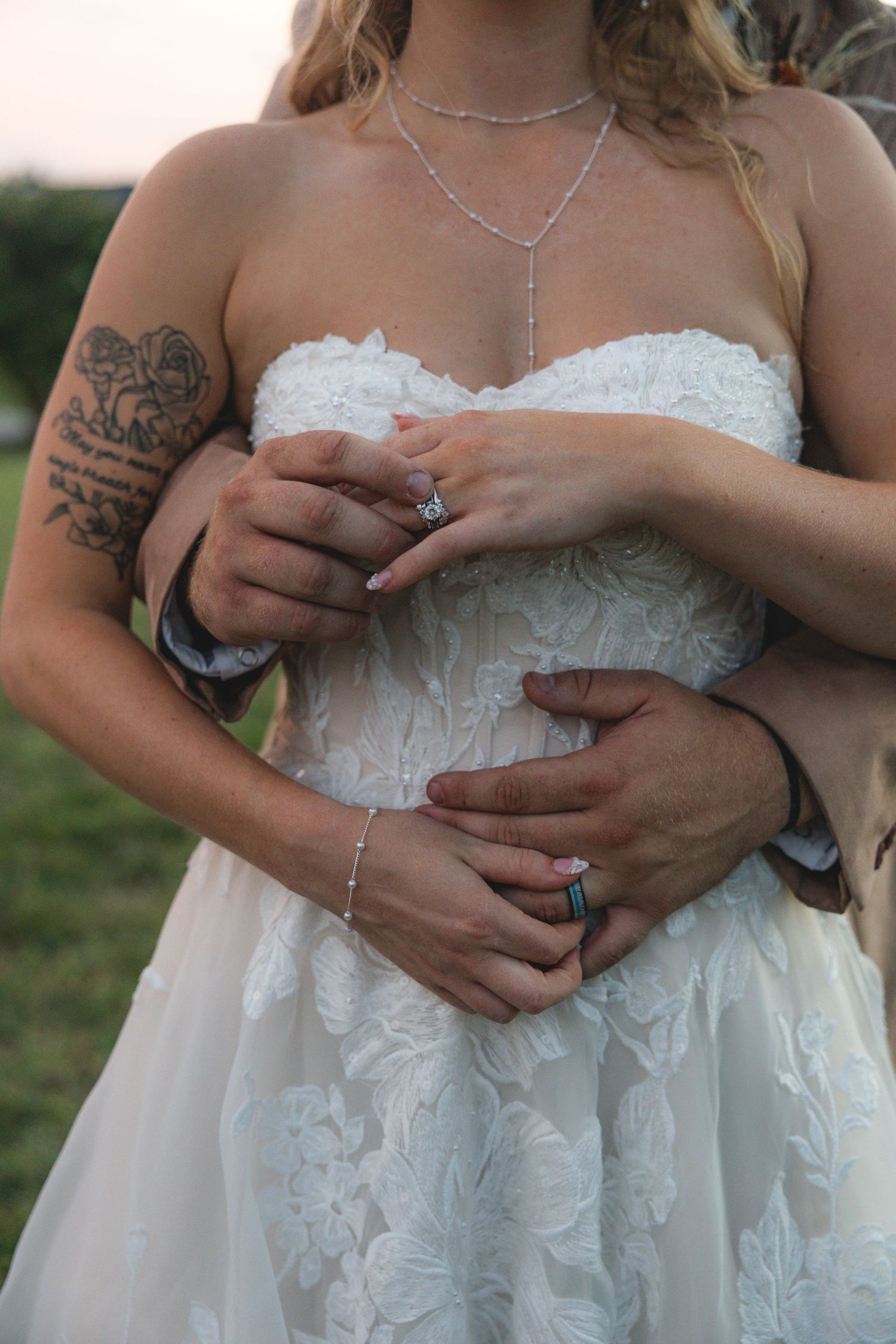 Bride in white lace strapless gown with tattoo, held by groom, both wearing wedding rings outdoors.