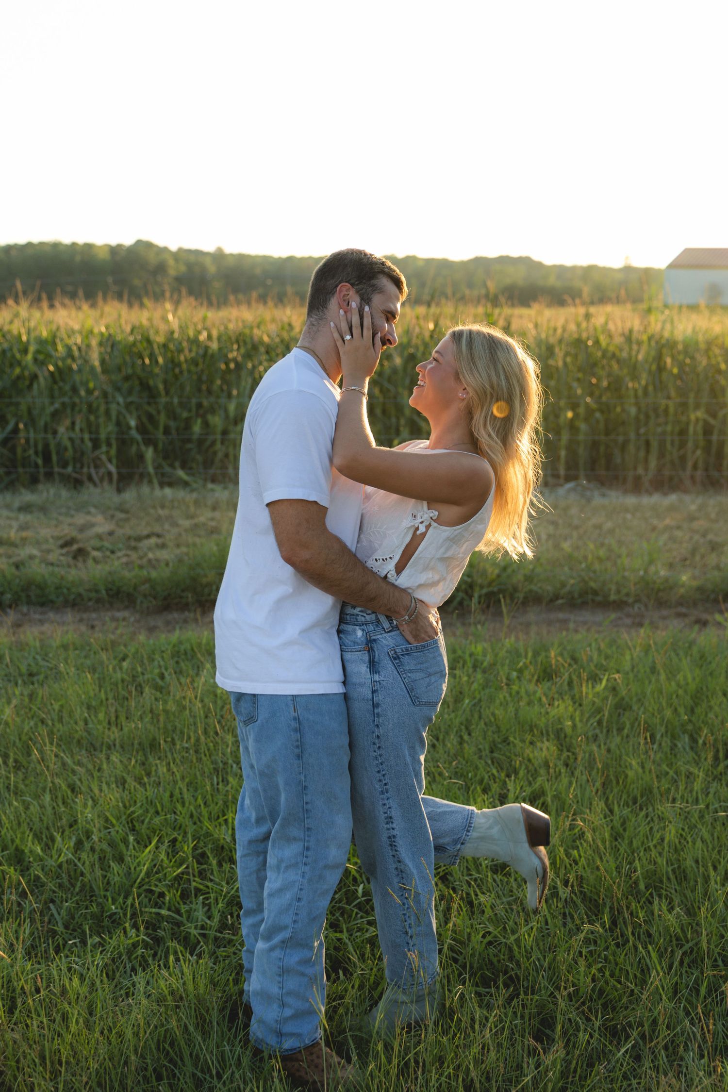Couple embracing and smiling at each other in a green field with a corn crop backdrop during golden hour.