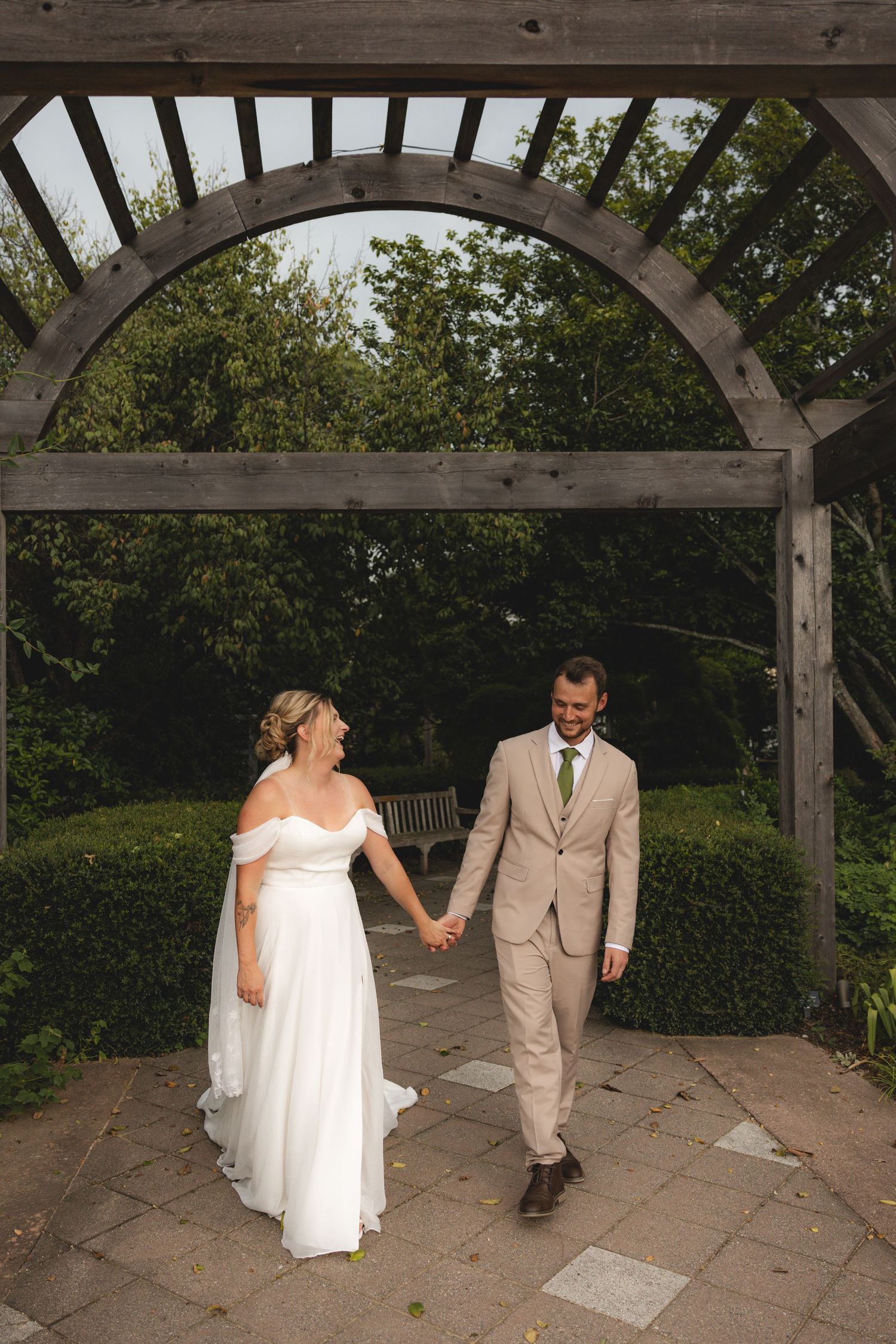 Bride and groom hold hands under a wooden arch in a lush garden, bride in white gown, groom in tan suit.