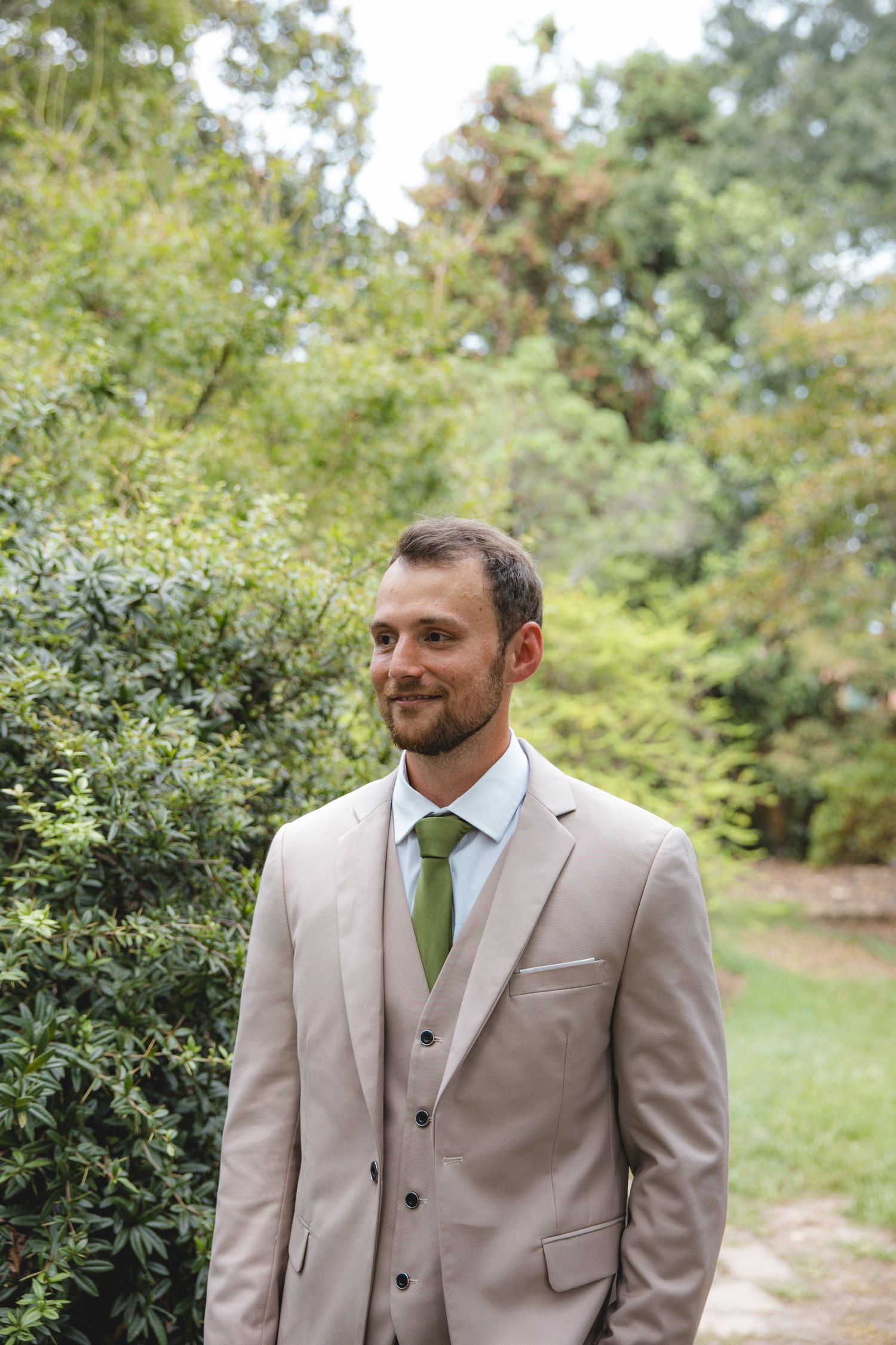 A smiling groom in a beige suit and green tie stands outdoors surrounded by lush green trees and shrubs.