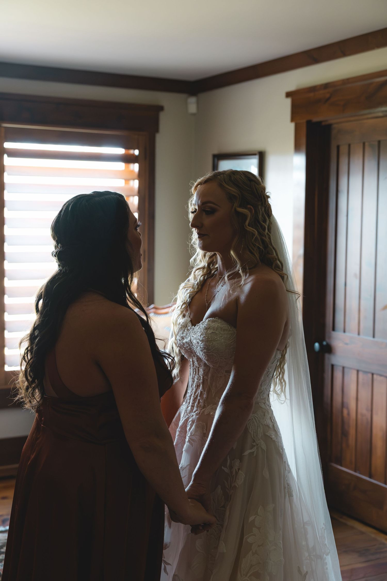 Bride in lace wedding gown and veil holds hands with bridesmaid in dark dress while getting ready indoors.