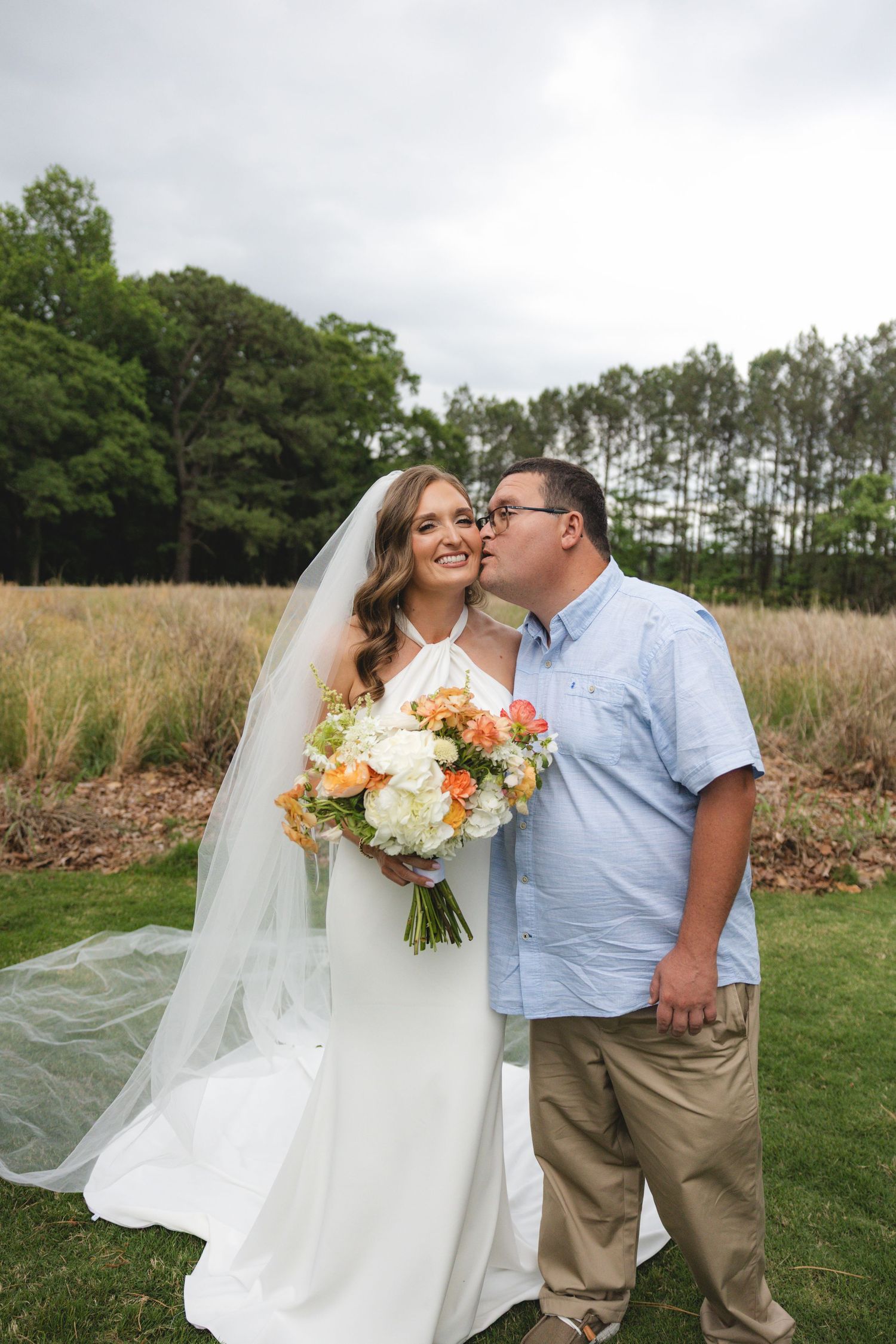 Bride in white gown with veil holds peach bouquet as groom in blue shirt kisses her cheek outdoors.