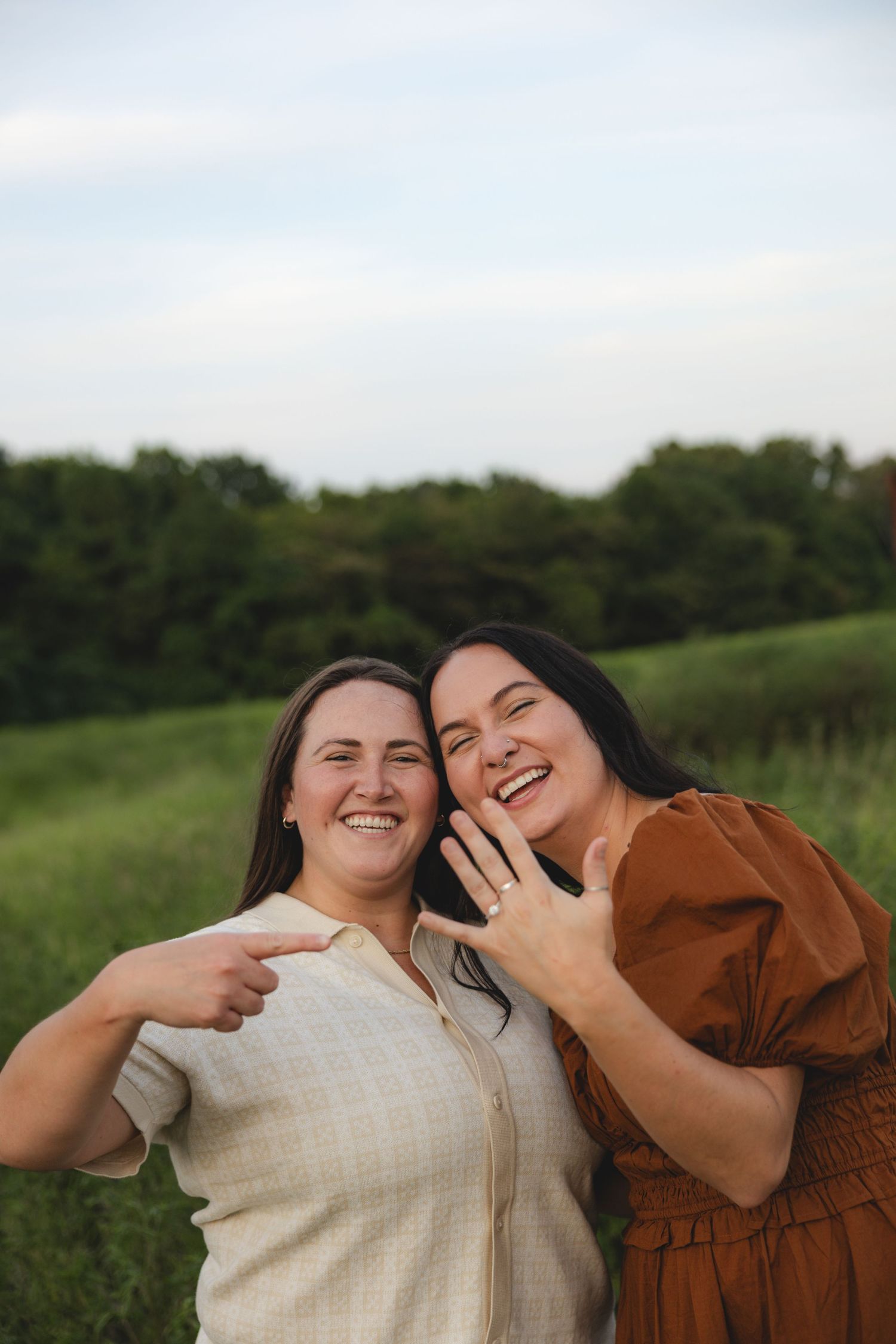 Two women smile joyfully outdoors, one pointing at the other's engagement ring in a green field.