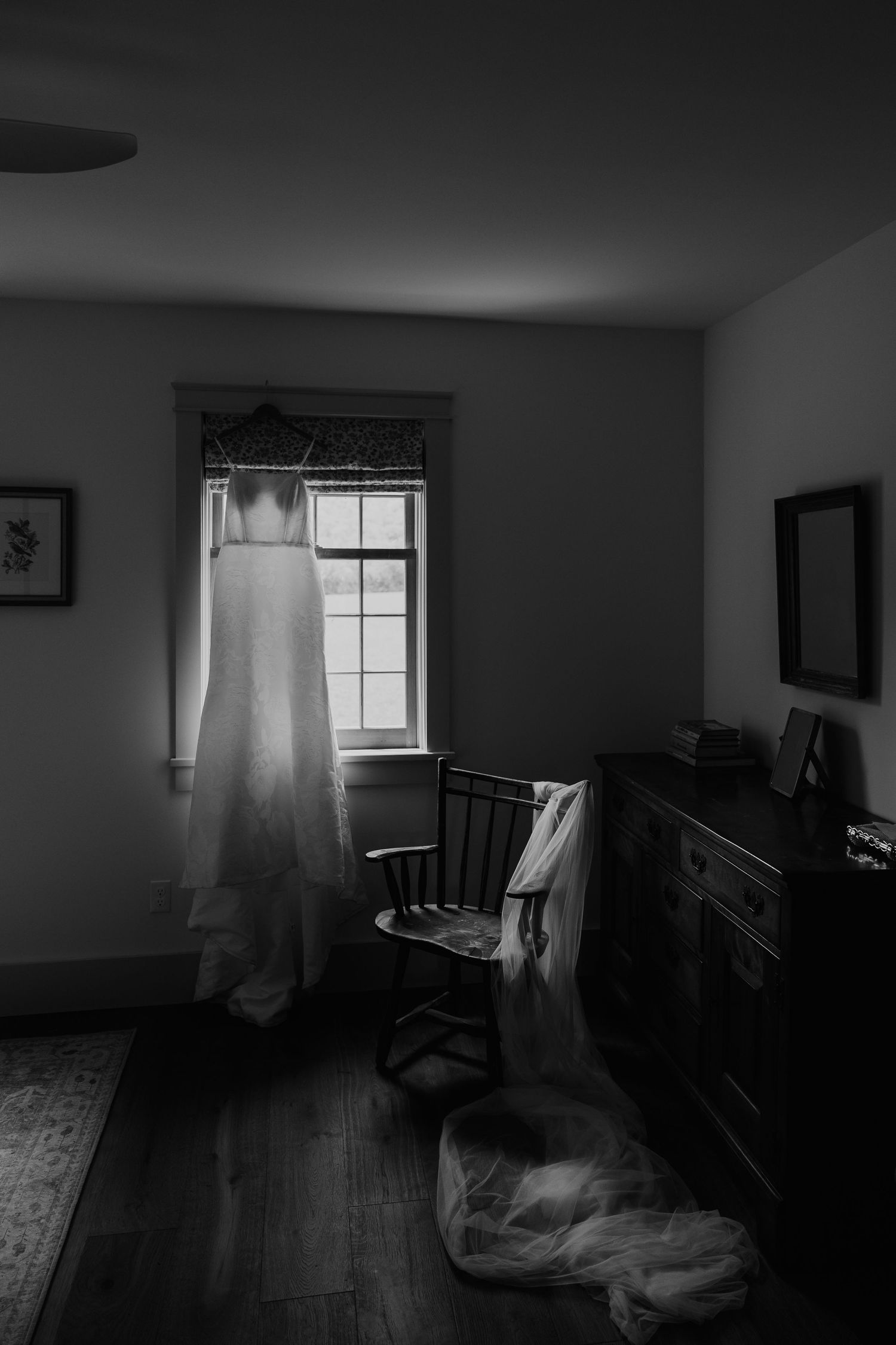 Black and white photo of a wedding dress hanging in a dimly lit room with a chair and dresser nearby.