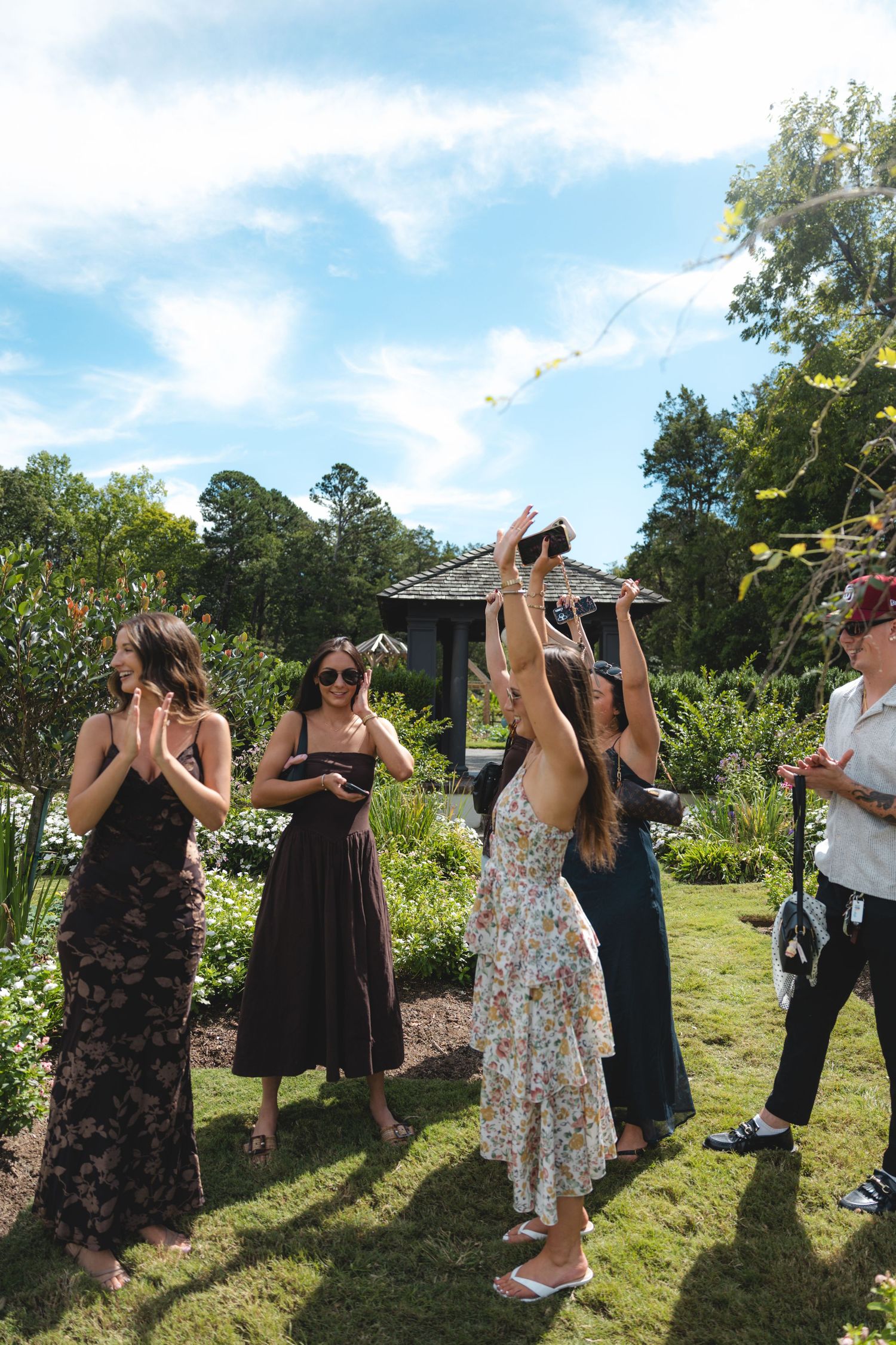 Women in elegant dresses gather outdoors in a garden, one reaching up to catch a bouquet at a sunny wedding celebration.