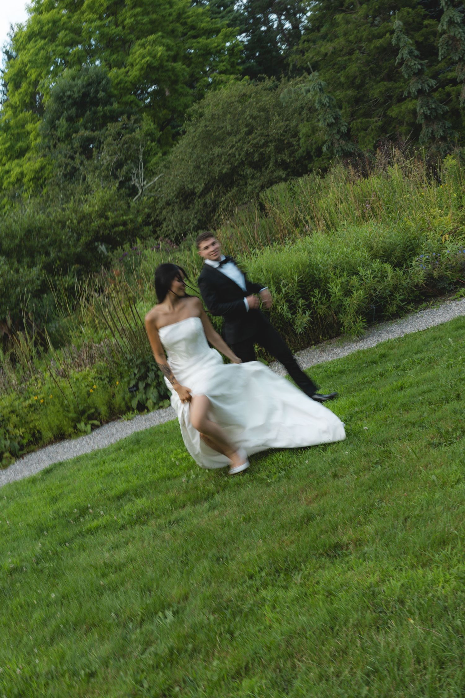 Bride in white strapless gown and groom in black tuxedo running together on green grass at outdoor wedding venue.
