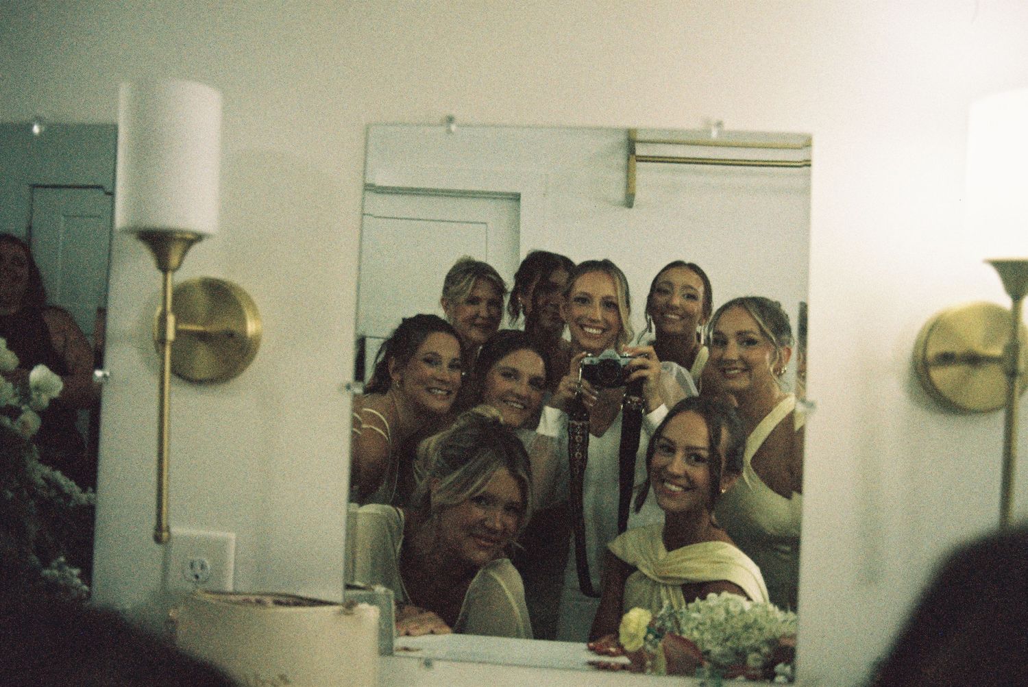 A group of smiling bridesmaids and wedding party take a mirror selfie in a bathroom with gold sconce lighting.