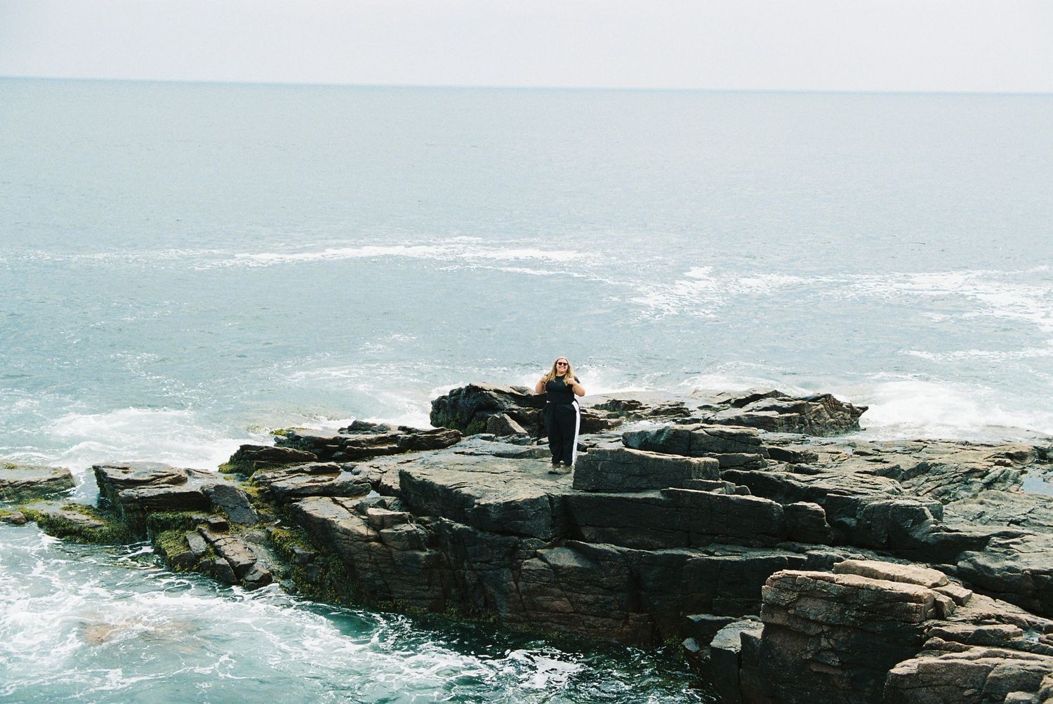 A lone figure stands on rugged coastal rocks overlooking the vast blue ocean waves on a misty day.
