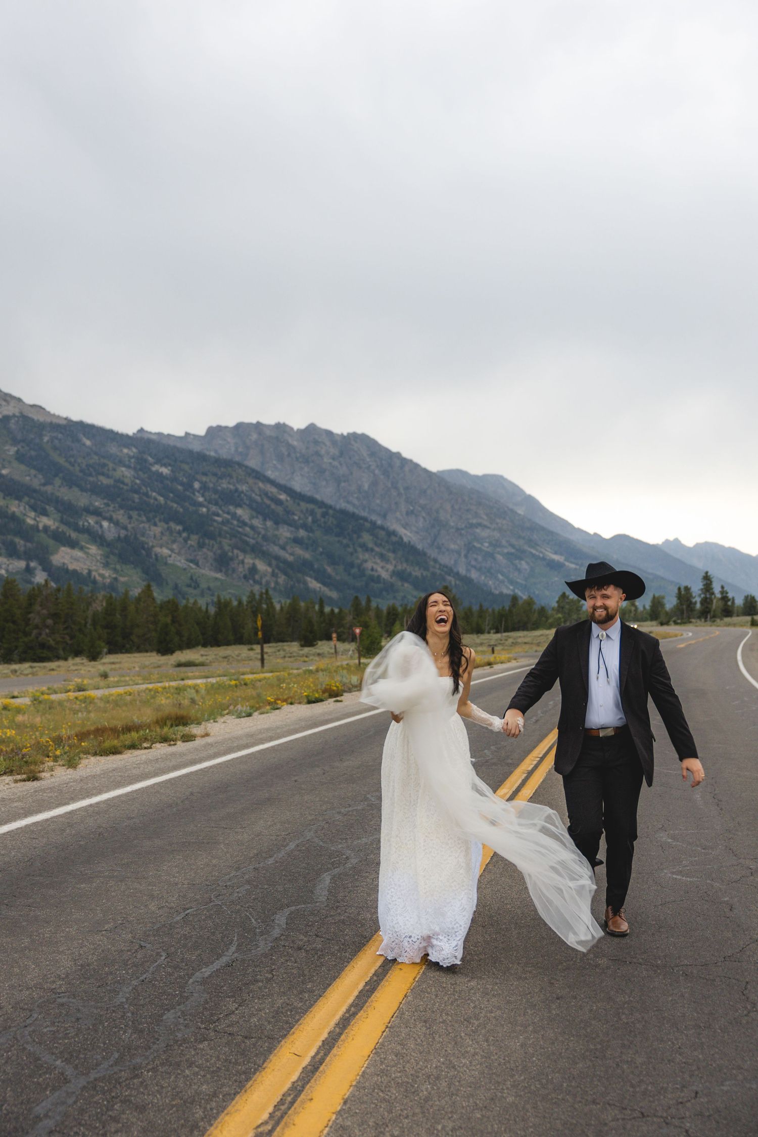Joyful bride and groom holding hands walking on mountain road, bride in flowing white dress, groom in black suit and cowboy hat.