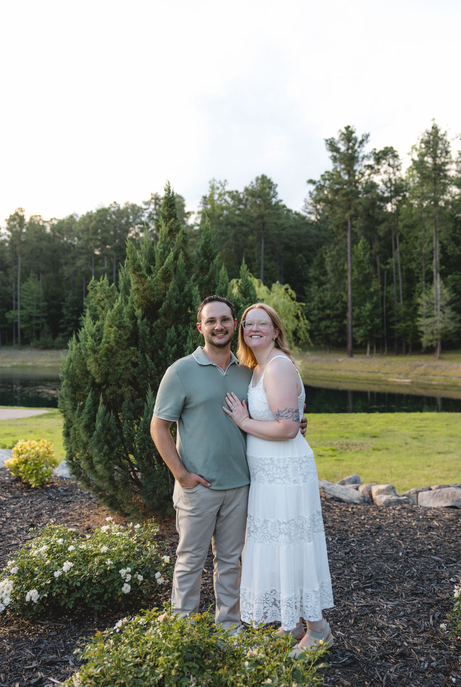 A smiling couple poses outdoors near evergreen trees and flowers at golden hour, woman in white dress, man in sage polo.