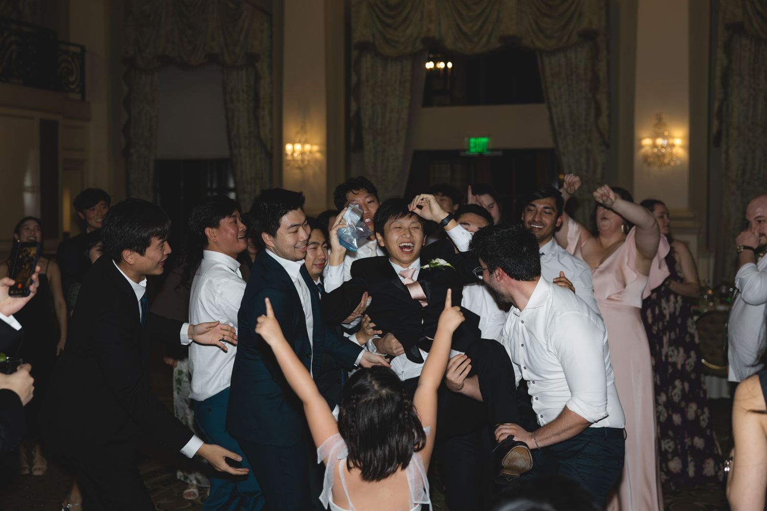 Energetic crowd of wedding guests dancing together in a grand ballroom during reception celebration.