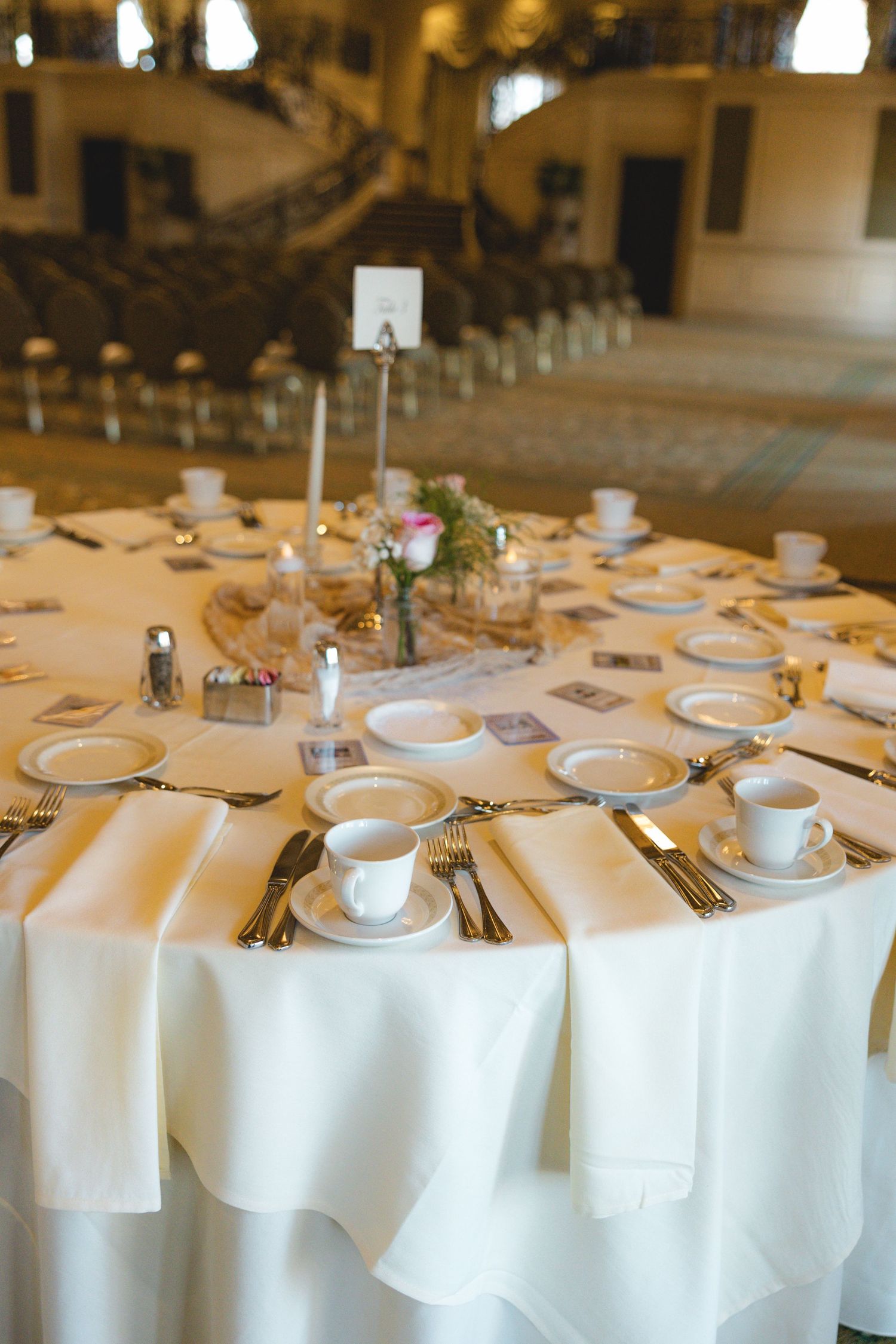 Formal wedding banquet table set with white linens, fine china teacups, gold cutlery, and floral centerpiece in grand hall.