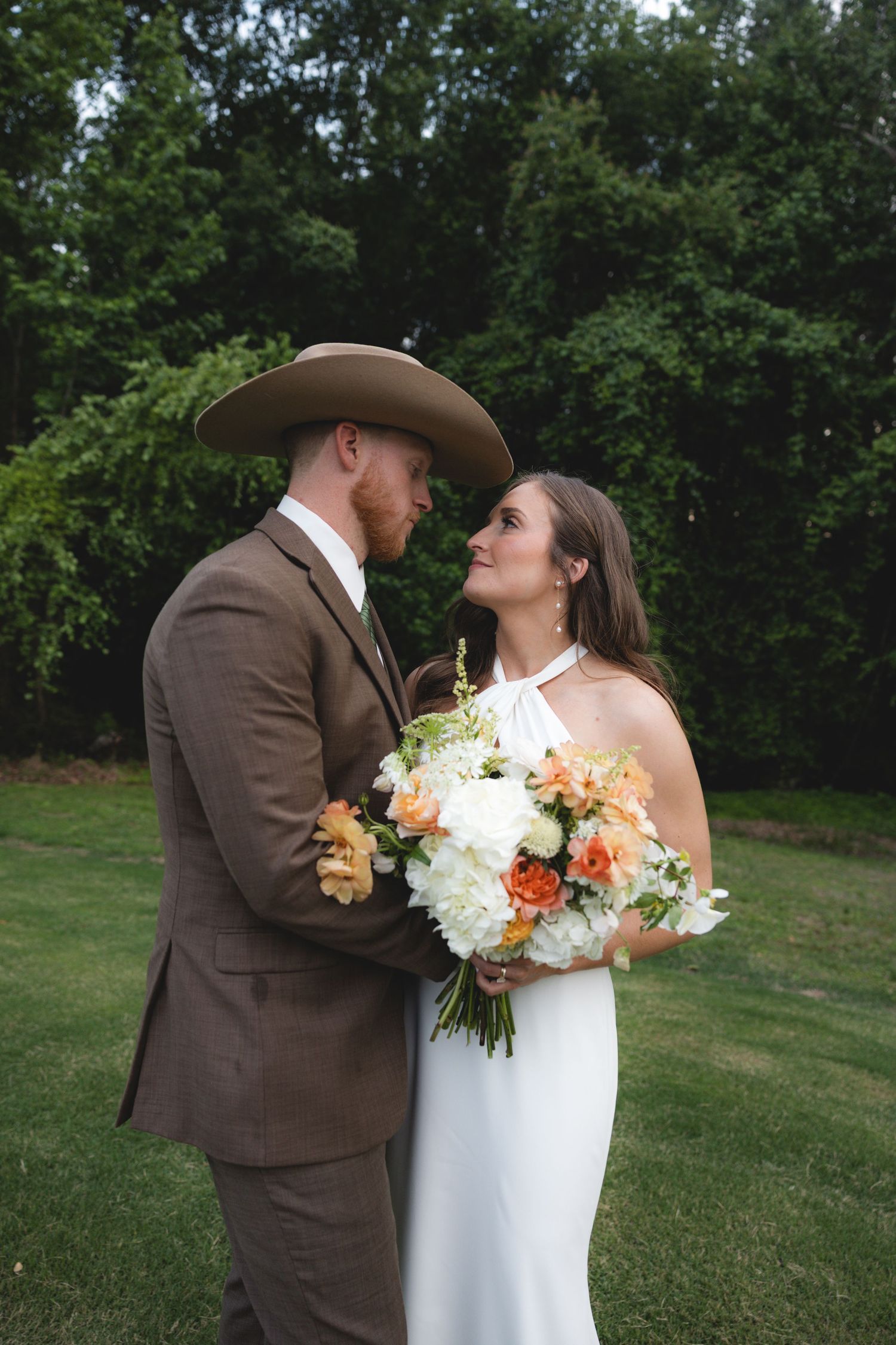 Bride and groom gazing at each other outdoors, groom in cowboy hat, bride holding peach and white bouquet.