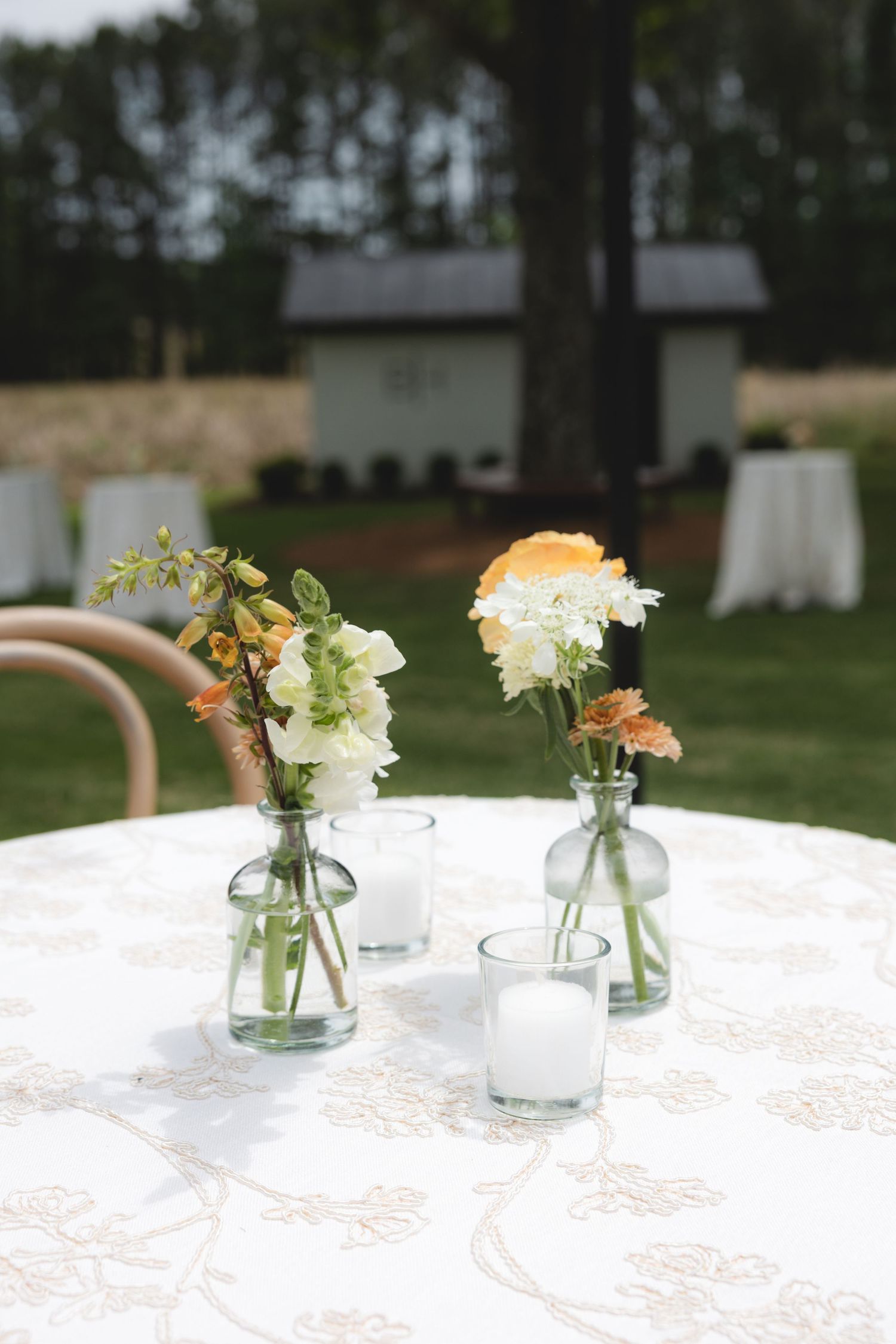 Rustic wedding table centerpiece with small glass bud vases holding white and peach flowers outdoors.