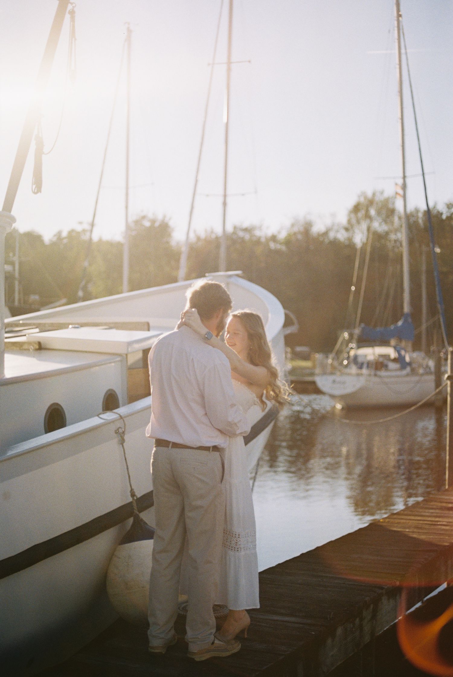 Couple embracing on a sunlit marina dock beside a sailboat during golden hour.