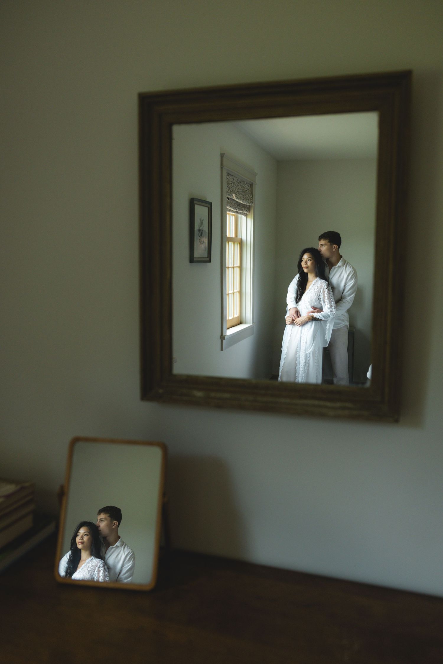 Couple in white robes embracing by a window, reflected in two mirrors inside a cozy, softly lit room.