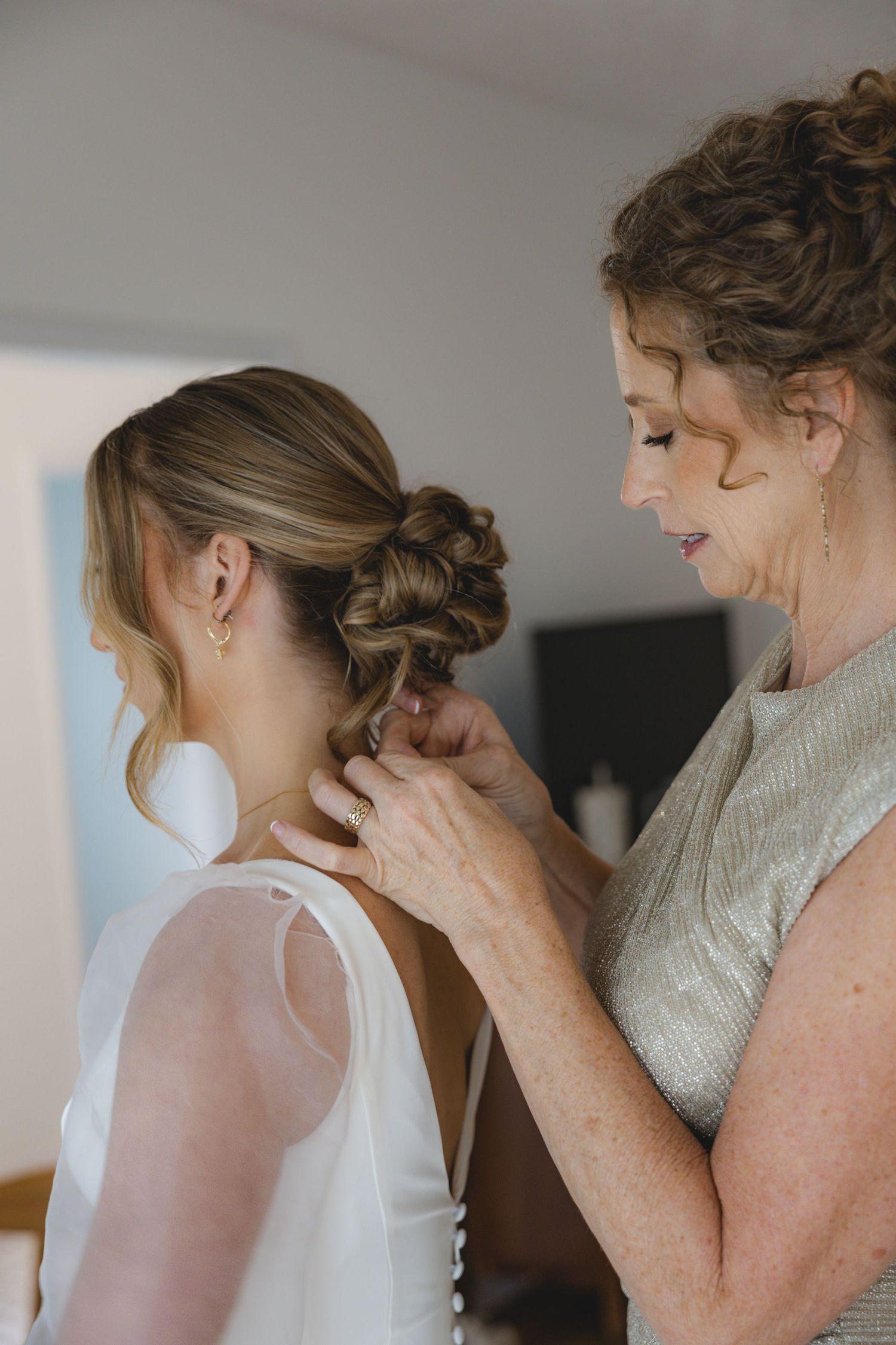A mother helps her daughter into a white wedding dress, adjusting the back while the bride's hair is styled in an elegant updo.