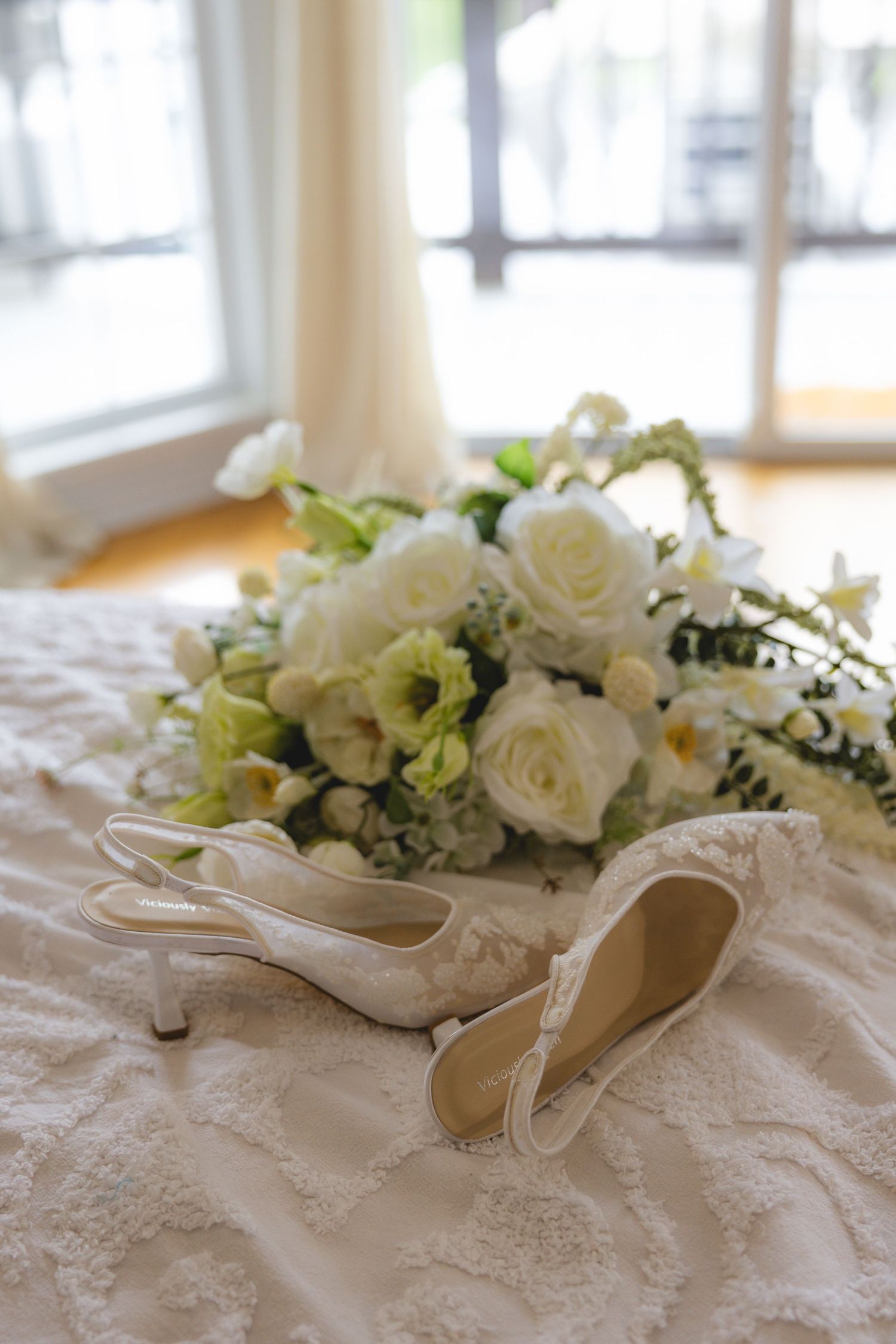 White lace bridal heels and elegant white rose bouquet arranged on a wedding dress near a sunlit window.
