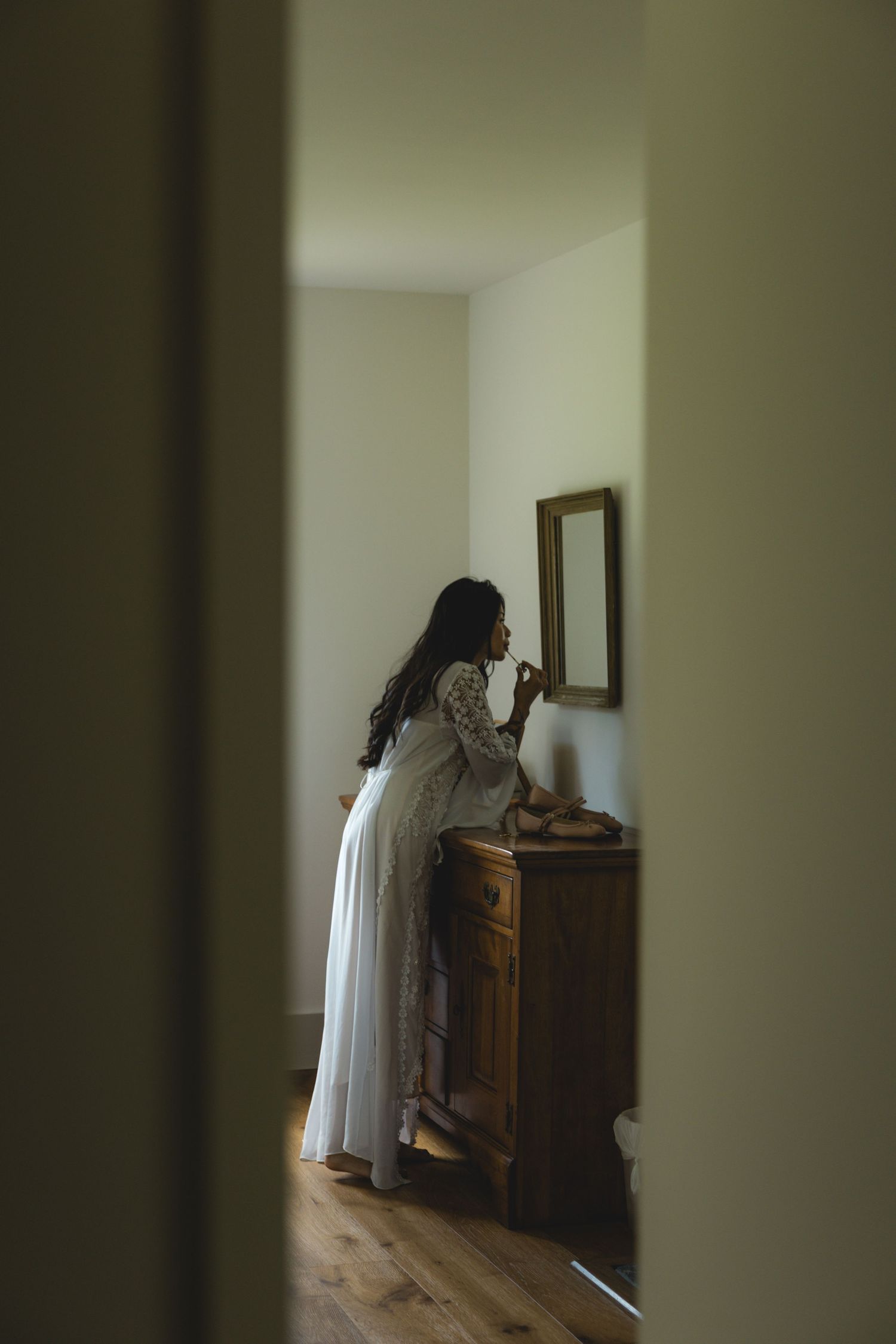 Bride in white lace gown standing at wooden dresser with mirror, preparing in softly lit room.