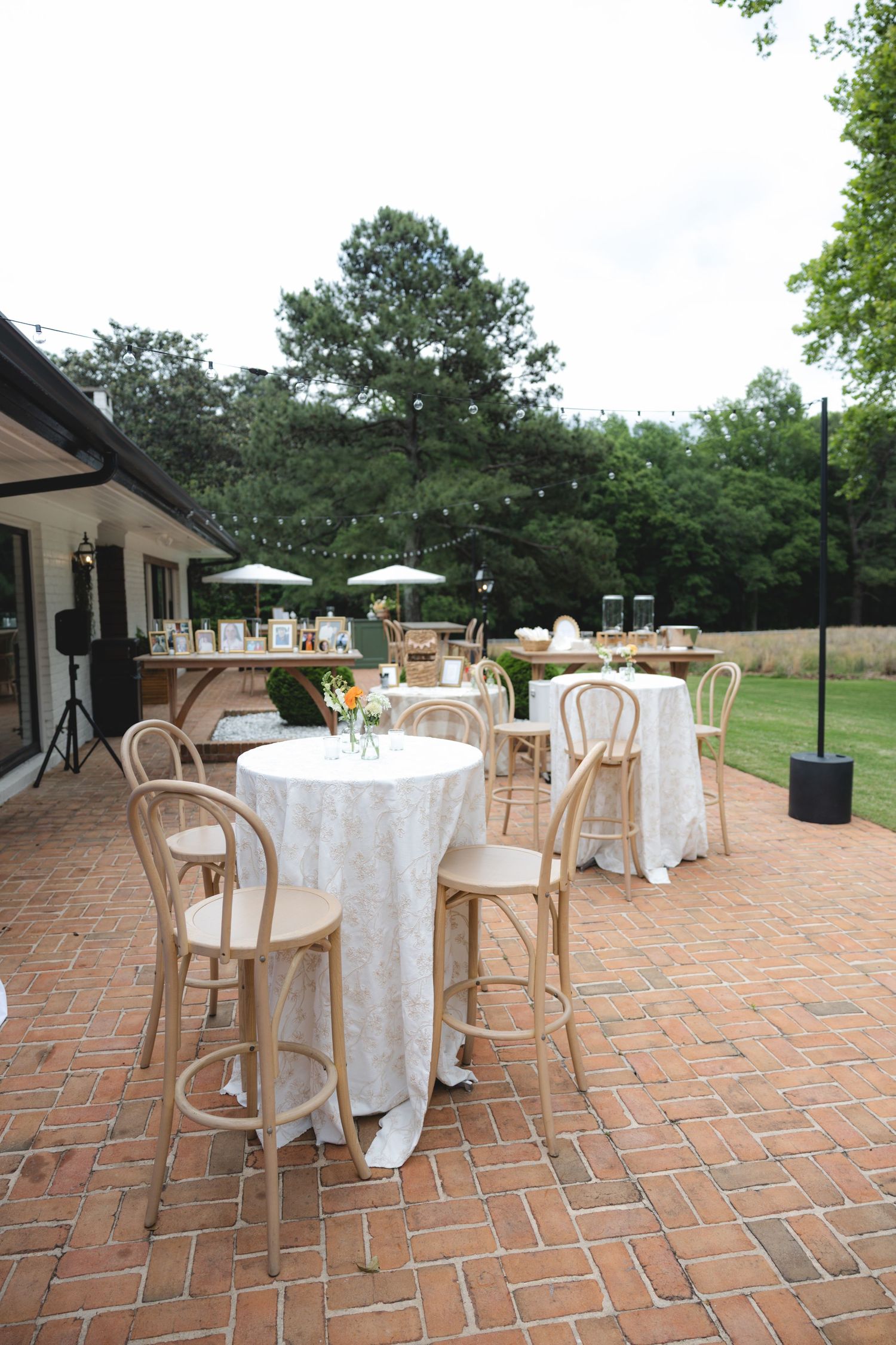 Elegant outdoor wedding reception on brick patio with white linen high-top tables, wooden bar stools, and lush green trees.