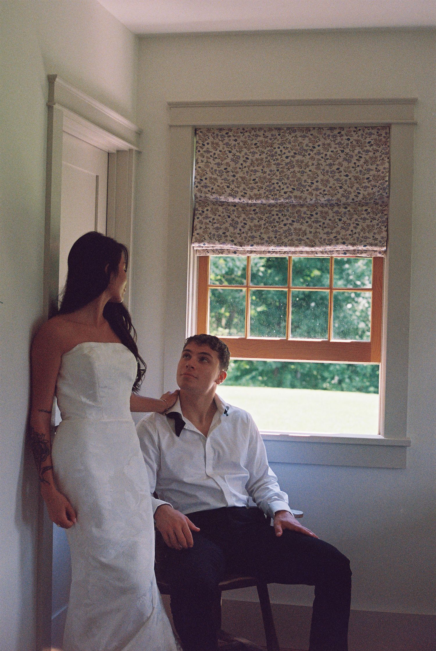 Bride in strapless white gown touches groom's shoulder as he sits by a window with floral roman shade.