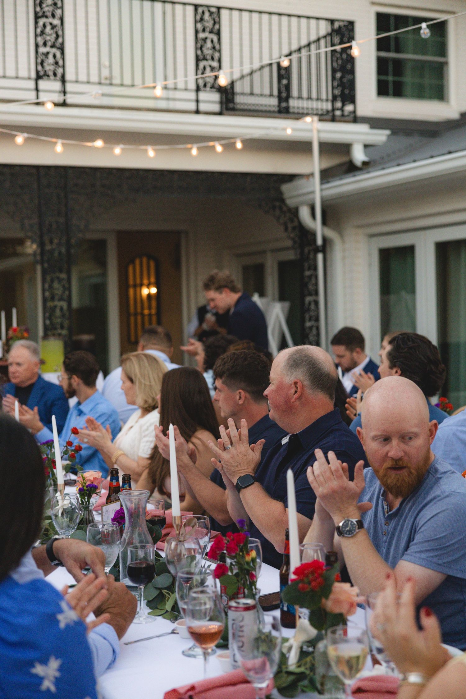 Guests clapping at an elegant outdoor dinner party with candles, wine glasses, and floral arrangements under string lights.