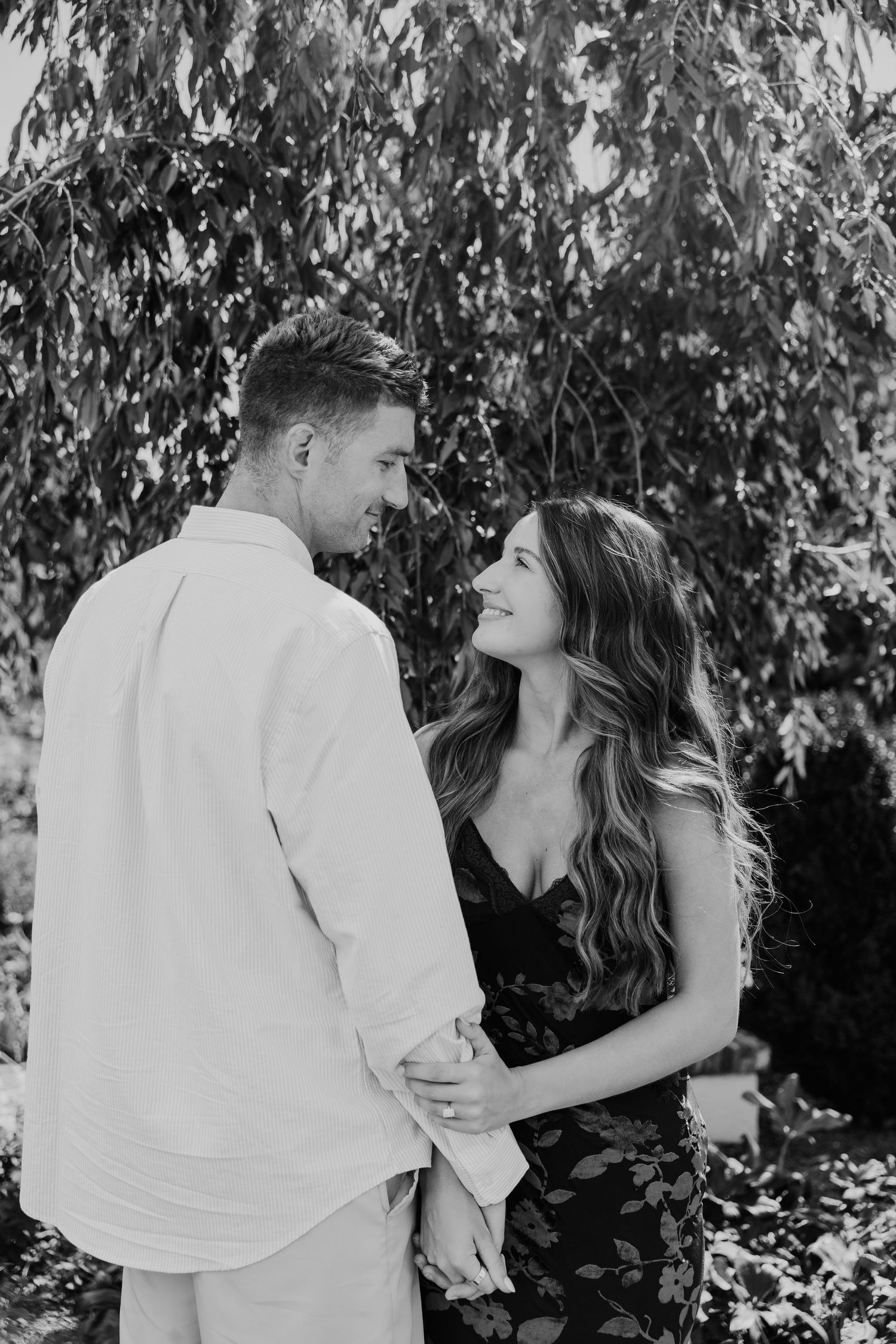 Black and white photo of a smiling couple holding hands and gazing at each other outdoors under lush tree branches.