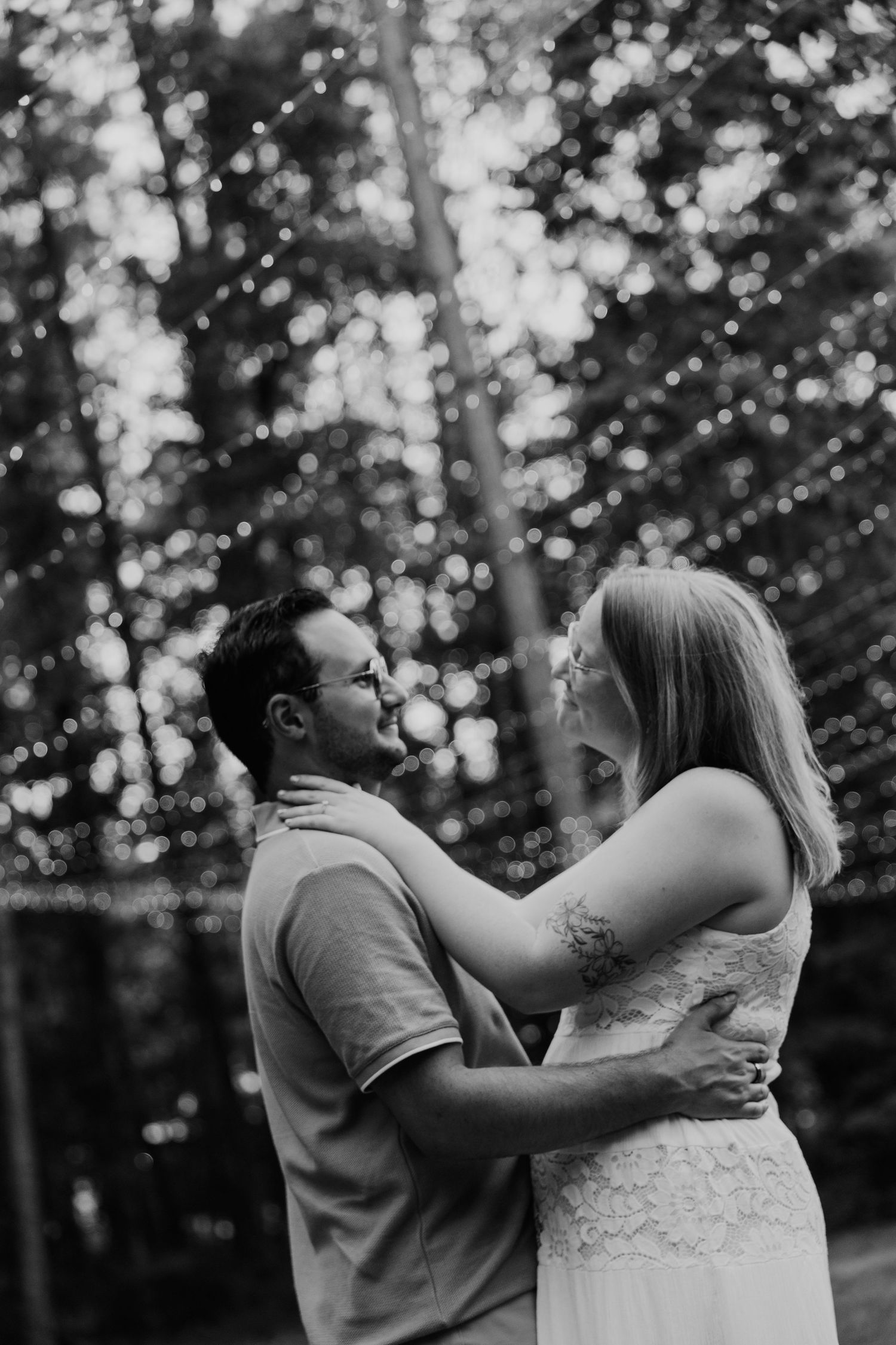 Black and white photo of a couple embracing outdoors with bokeh string lights and trees in the background.