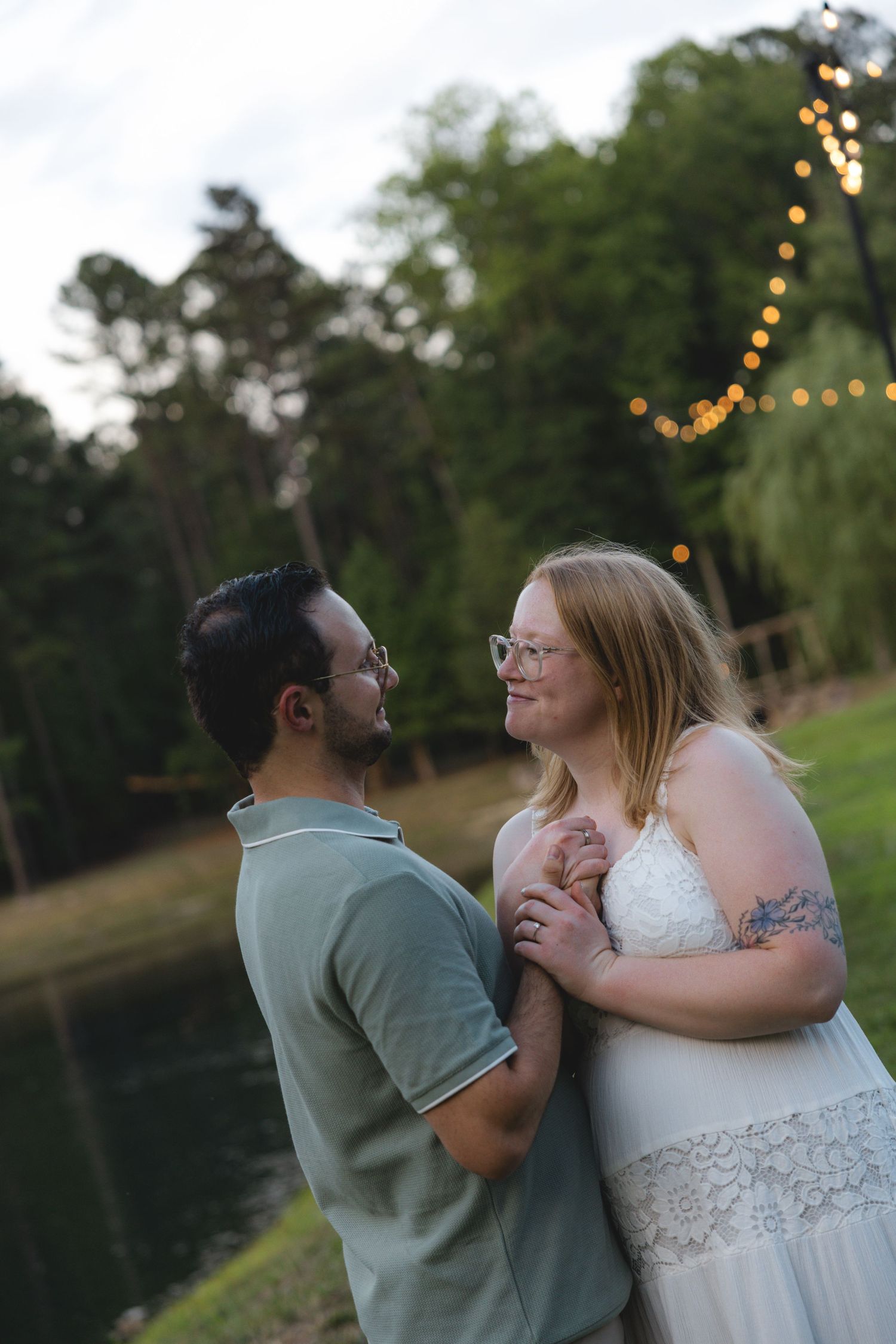 Couple sharing a romantic moment outdoors near a pond with string lights at dusk, woman in white lace dress.