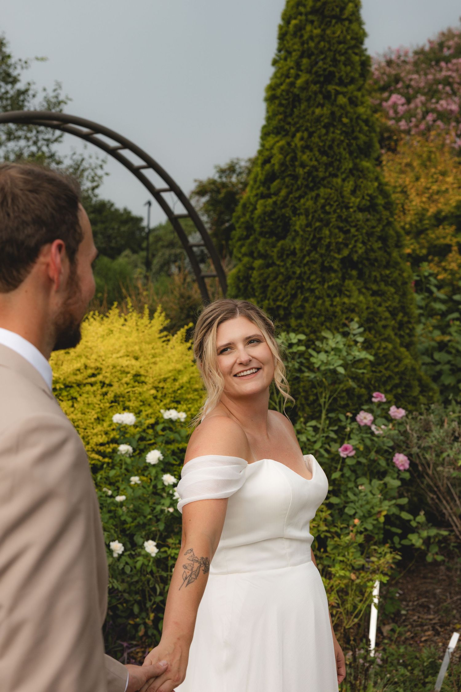 Smiling bride in off-shoulder white gown holds hands with groom in tan suit at outdoor garden wedding ceremony.