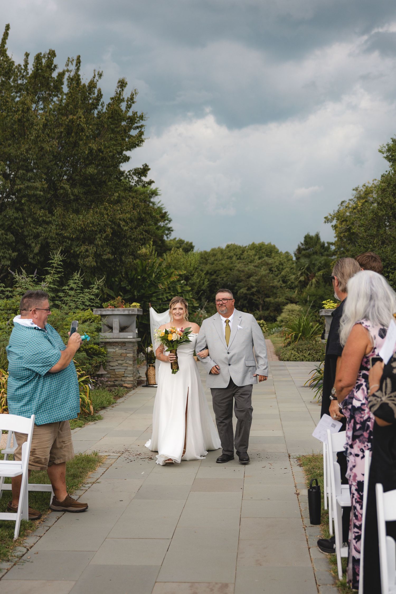 Bride walks down outdoor aisle with escort, guests watching, lush greenery and dramatic cloudy sky backdrop.