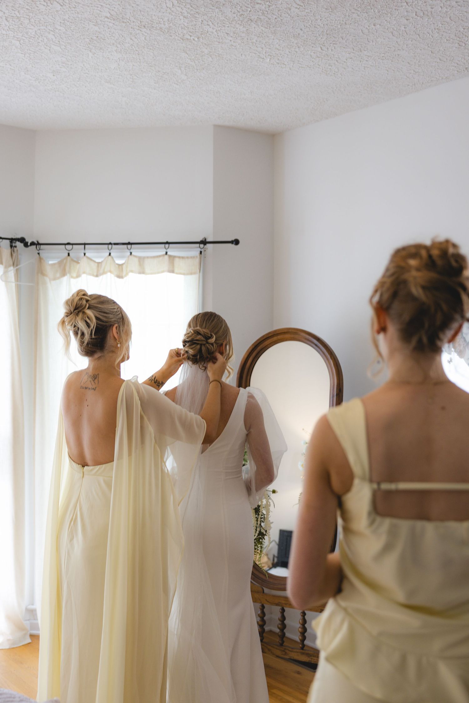 Bridesmaids helping bride with veil in front of oval mirror before wedding ceremony.