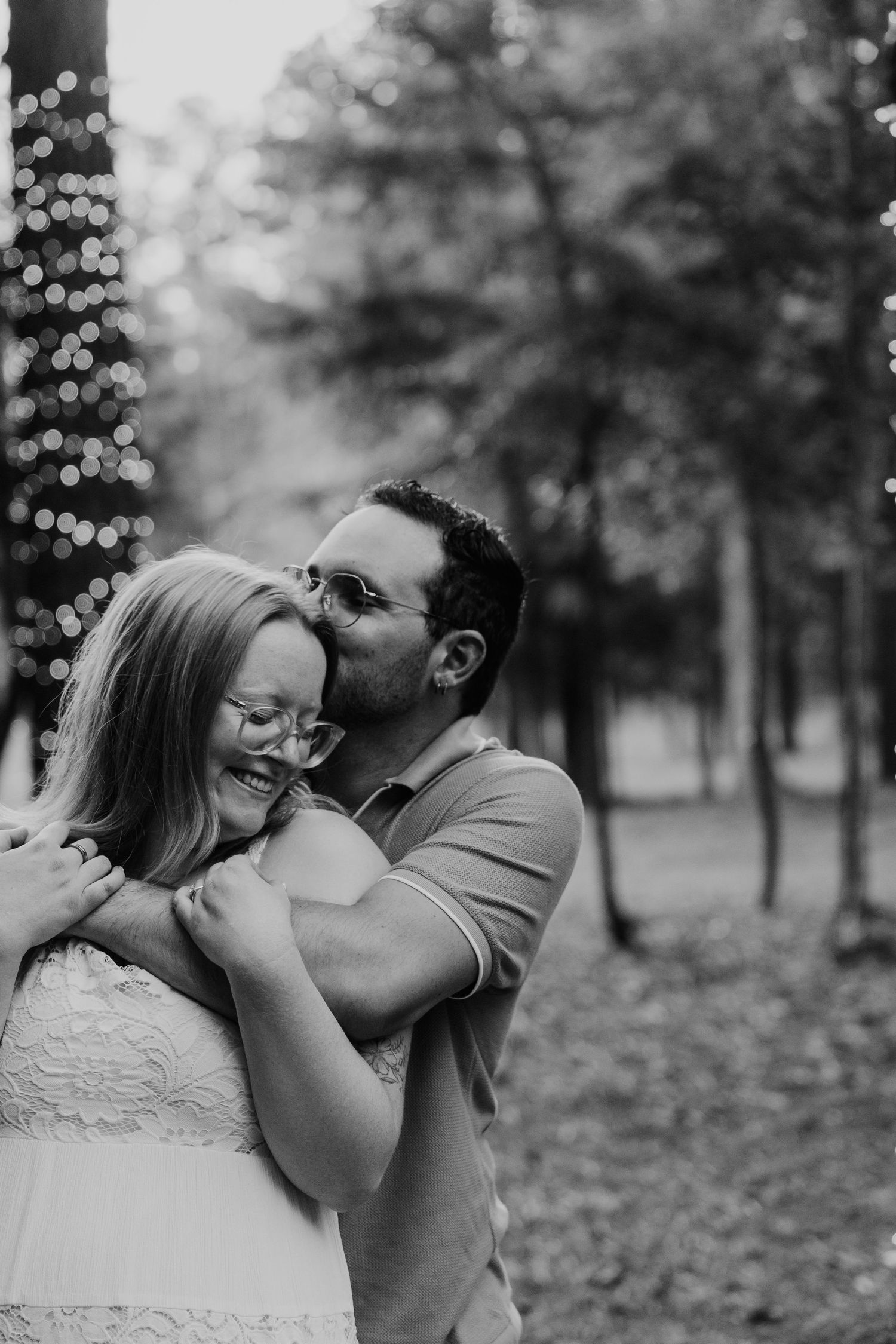 Black and white photo of a couple embracing outdoors, man kissing woman on cheek as she laughs among trees.