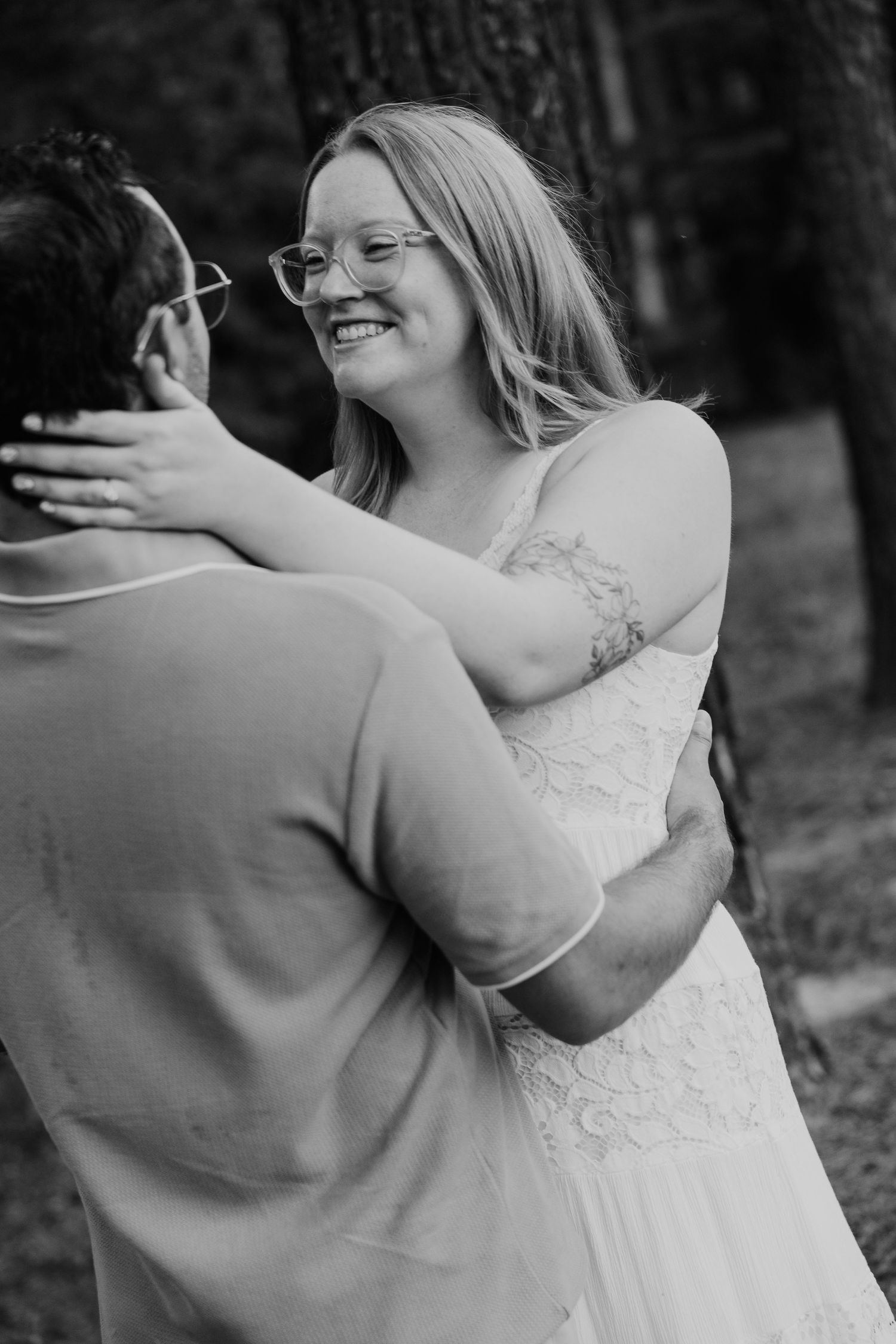 Black and white photo of a smiling bride in a lace dress embracing her partner outdoors.