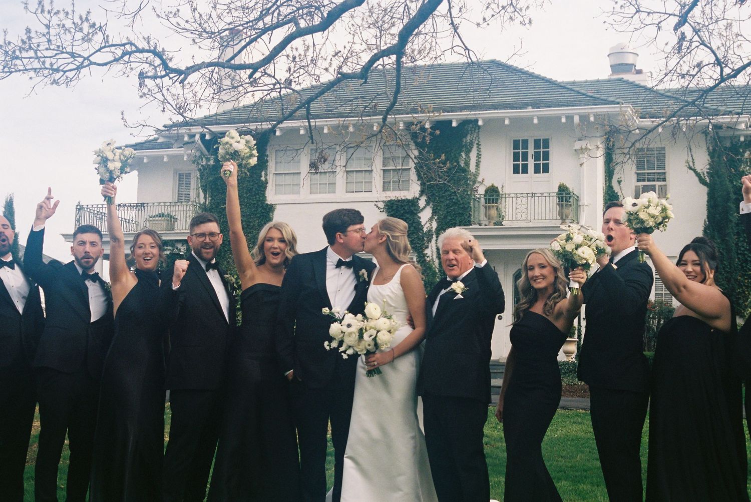 A wedding party gathered outdoors in front of a historic building, with the bride and groom standing in the center.