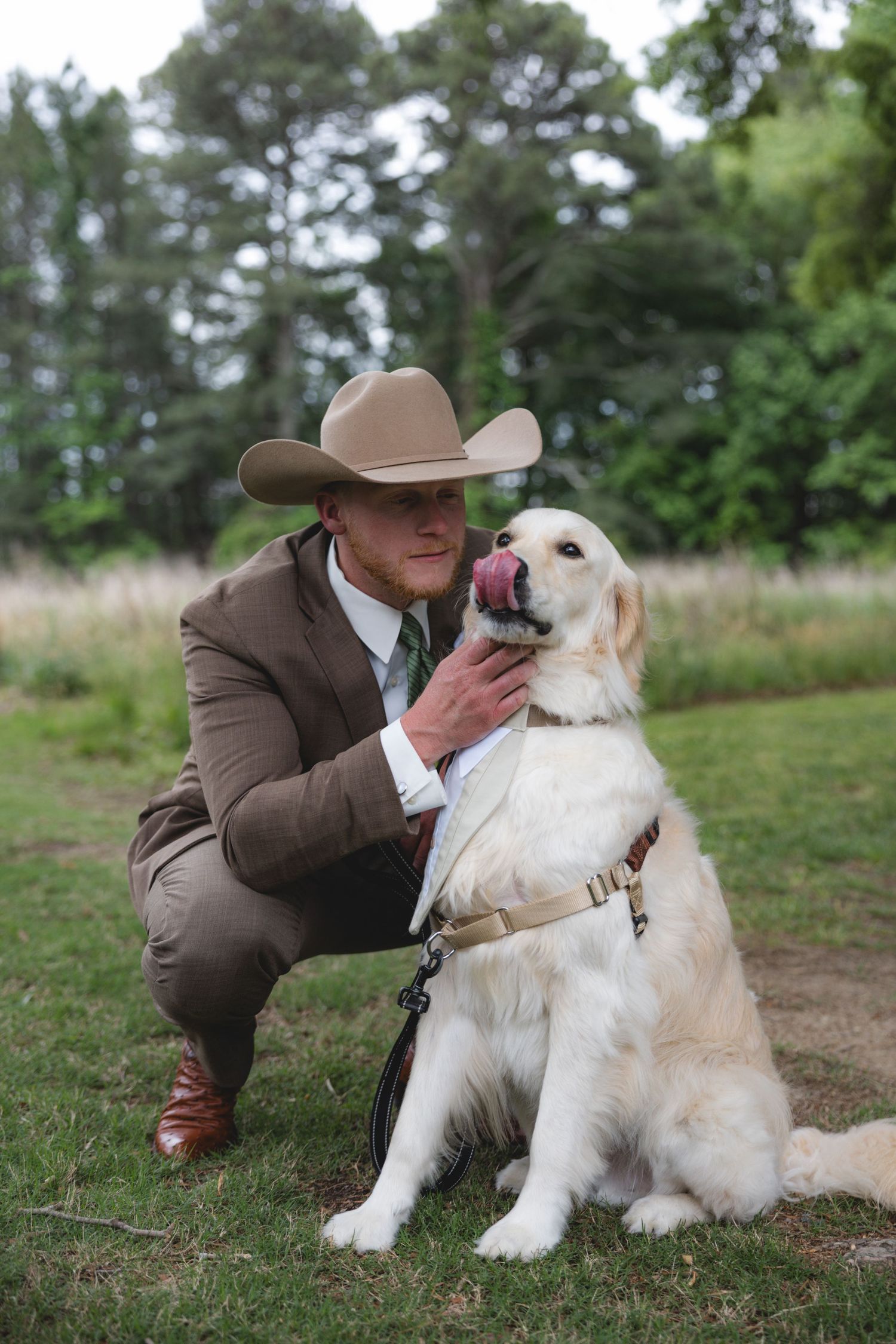 Man in brown suit and cowboy hat crouching beside a golden retriever on a leash in a grassy outdoor setting.
