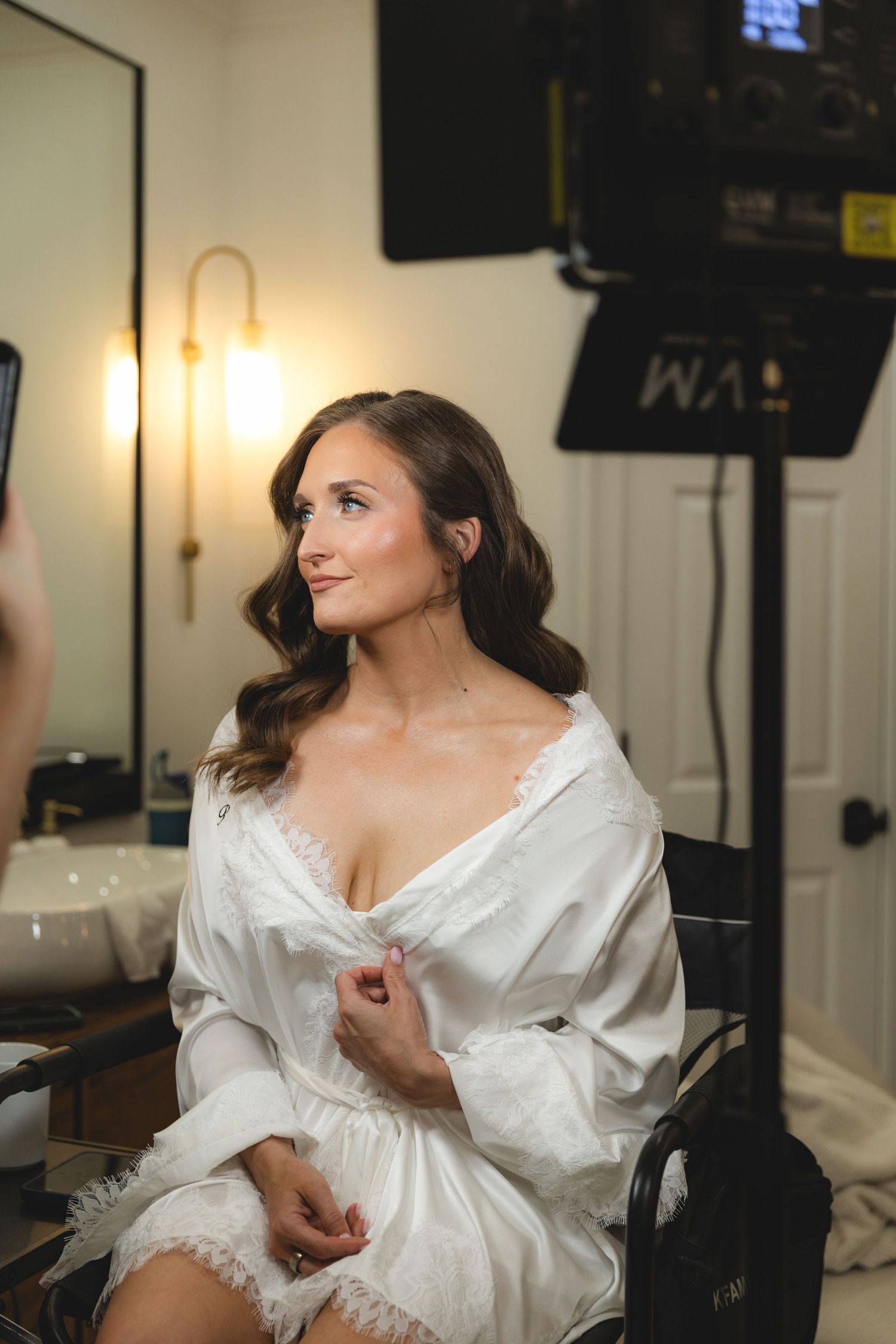 Bride in white lace robe getting ready in bridal suite with professional lighting and camera equipment.
