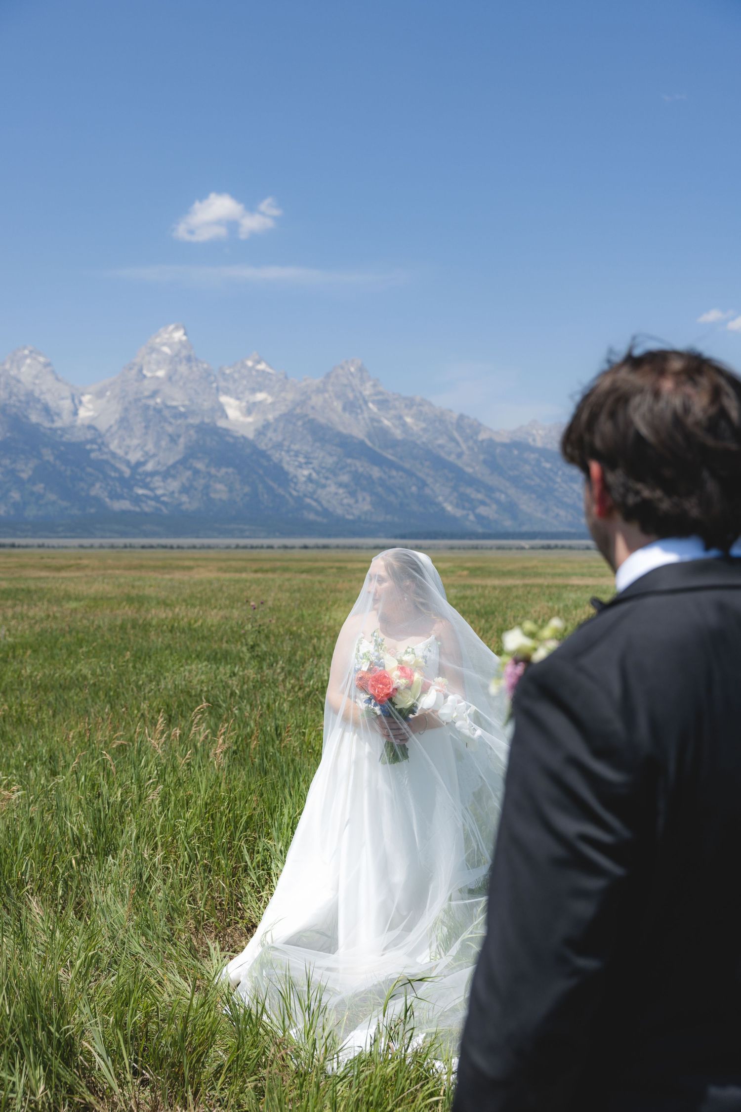 Bride in white gown and veil holds bouquet in green meadow with Grand Teton mountains backdrop on sunny wedding day.