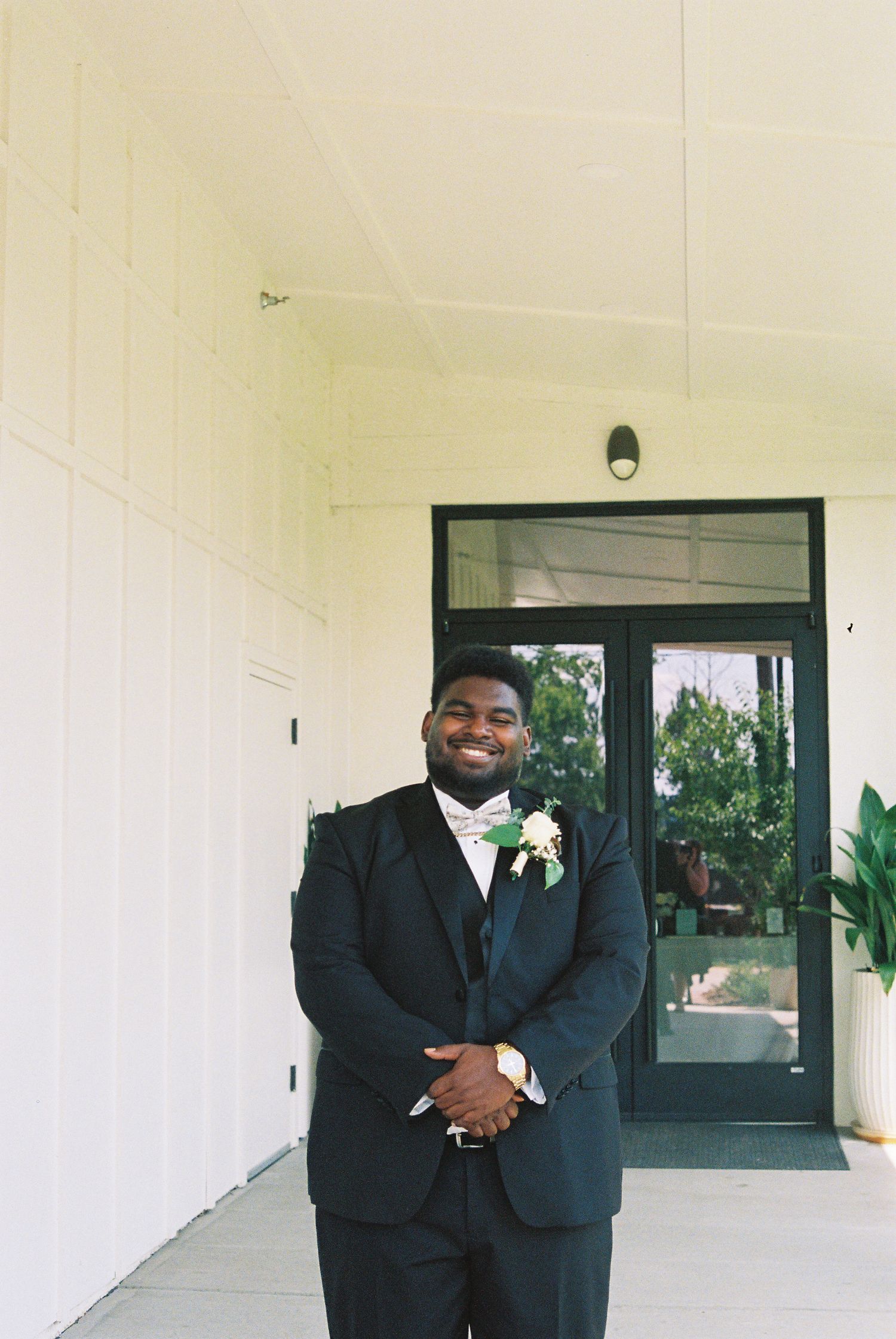 A smiling groom in a black tuxedo with bow tie and boutonniere stands outside a white building entrance.