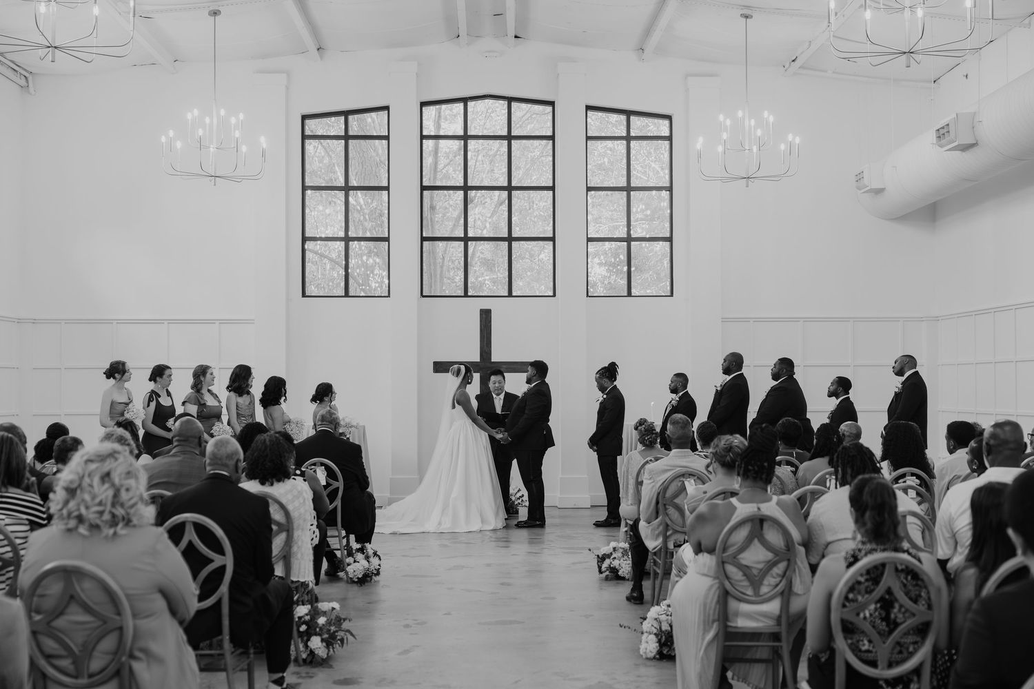 Black and white wedding ceremony photo in elegant white chapel with large windows, couple at altar with cross.