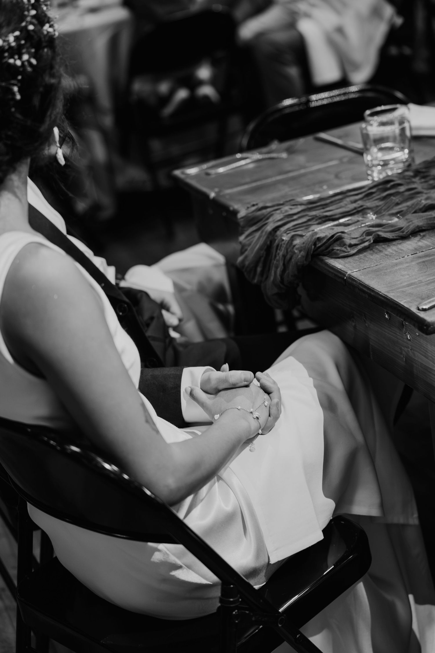 Black and white photo of a bride in white dress sitting at a wooden table with hands folded in her lap.