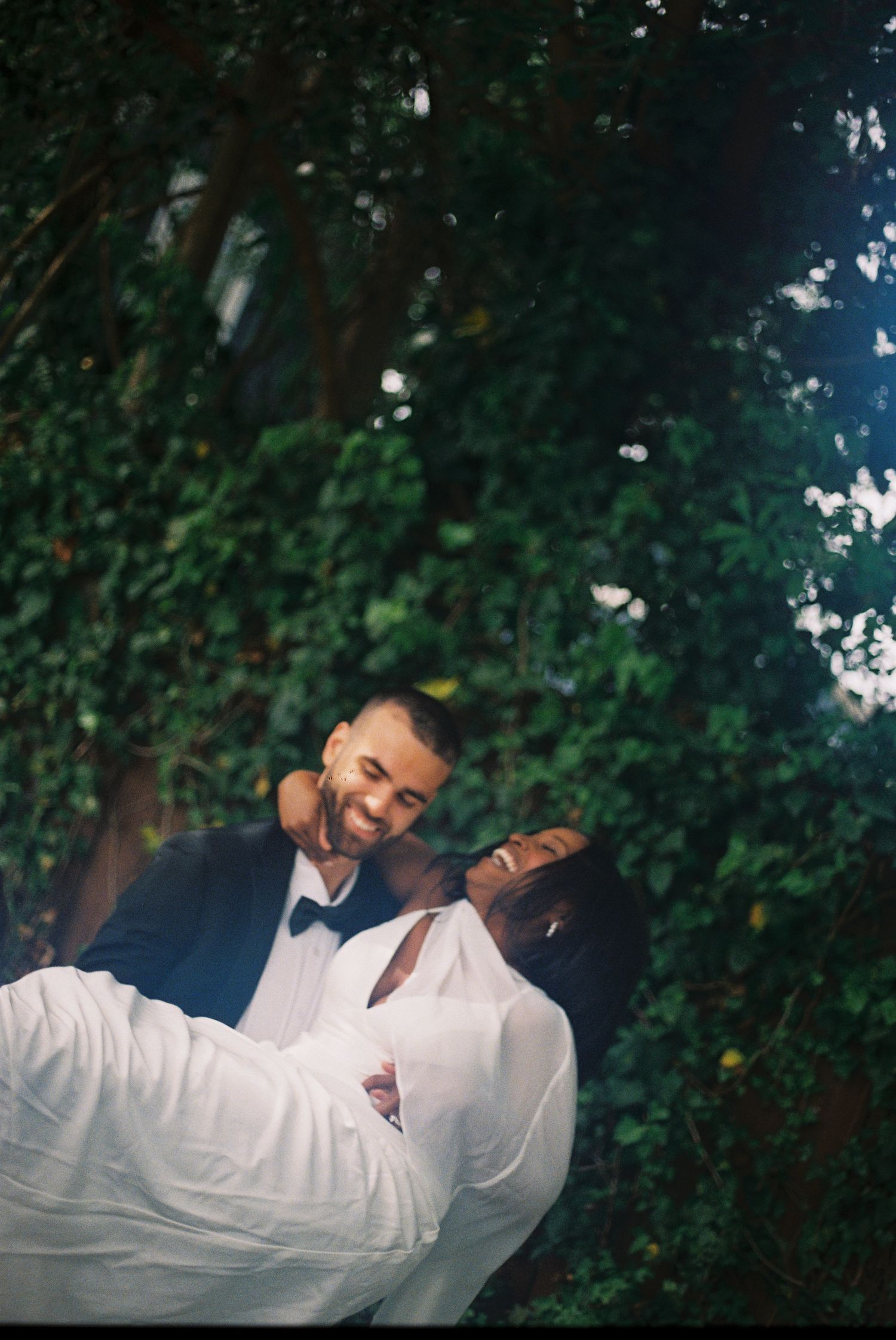 A groom in a navy suit carries his bride in a white dress, both smiling joyfully, surrounded by lush green foliage.