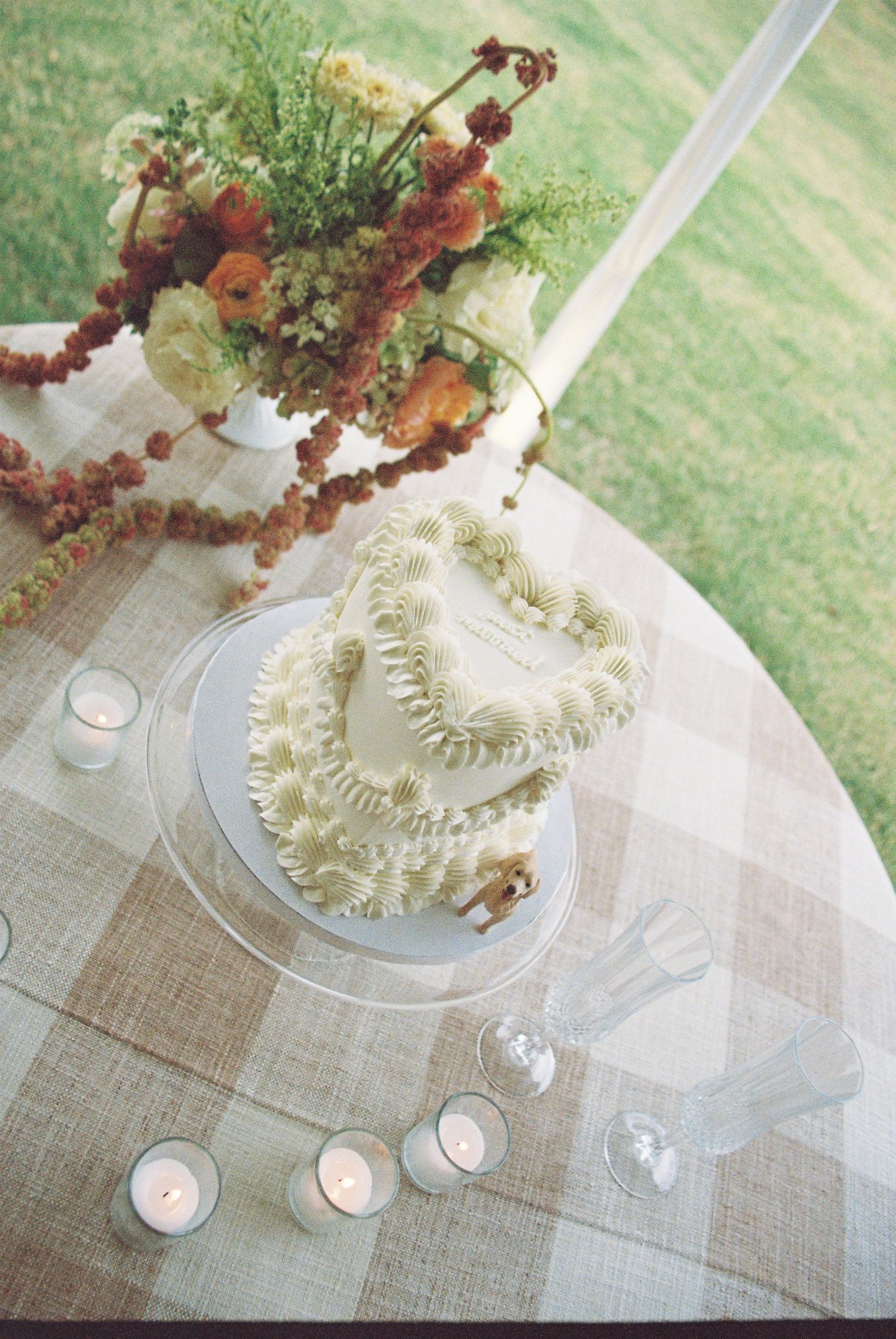 Heart-shaped white wedding cake with ruffled frosting on glass stand, surrounded by candles and floral arrangement.