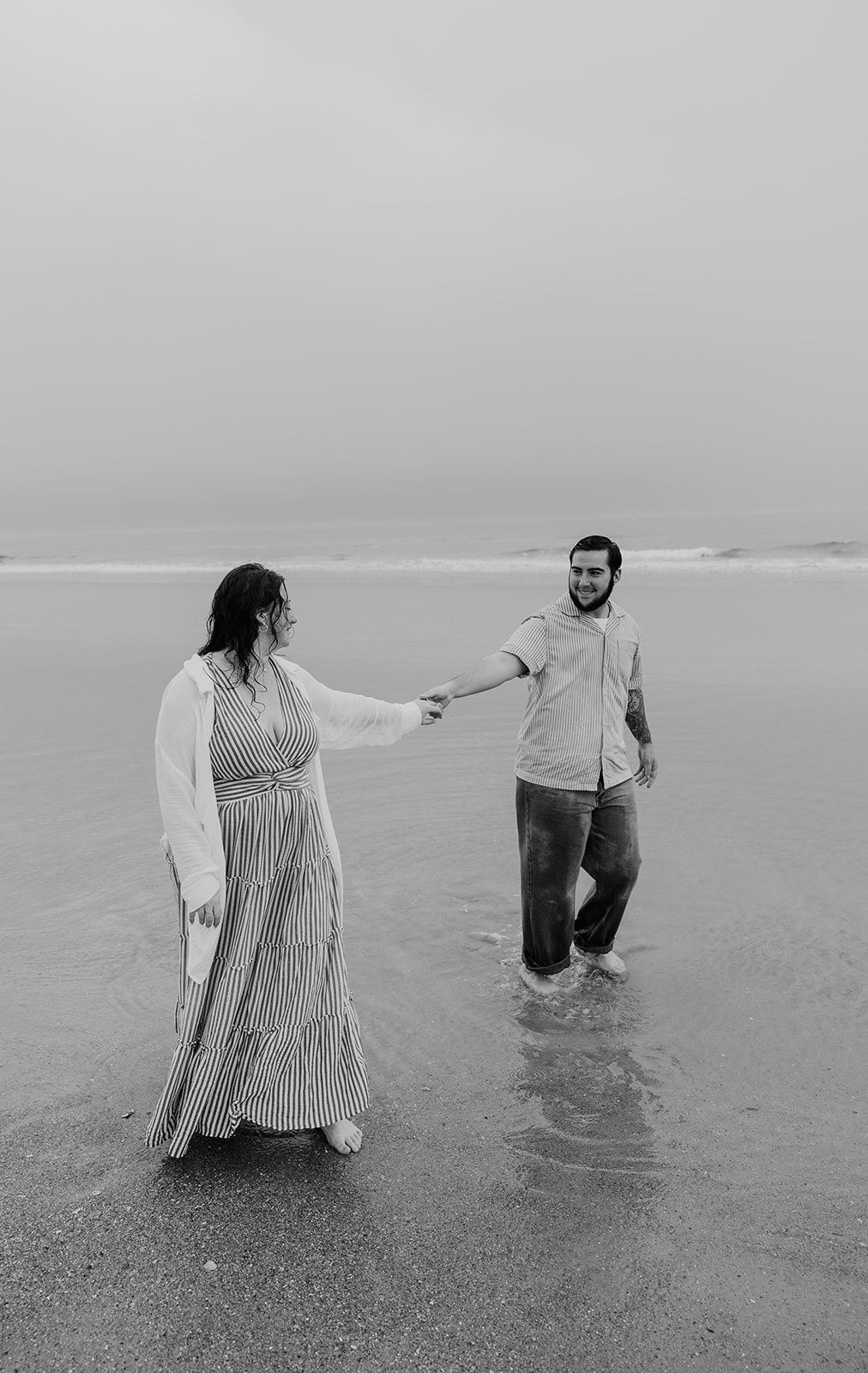 Black and white photo of a couple holding hands on a beach, standing in shallow water, smiling at each other.